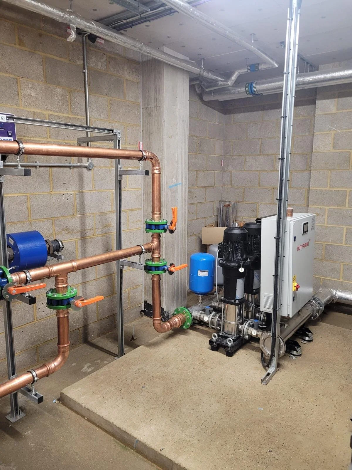 Industrial plumbing setup with copper pipes, valves, and a black pump. There are also a blue expansion tank, a gray control panel, and silver ductwork on the ceiling.