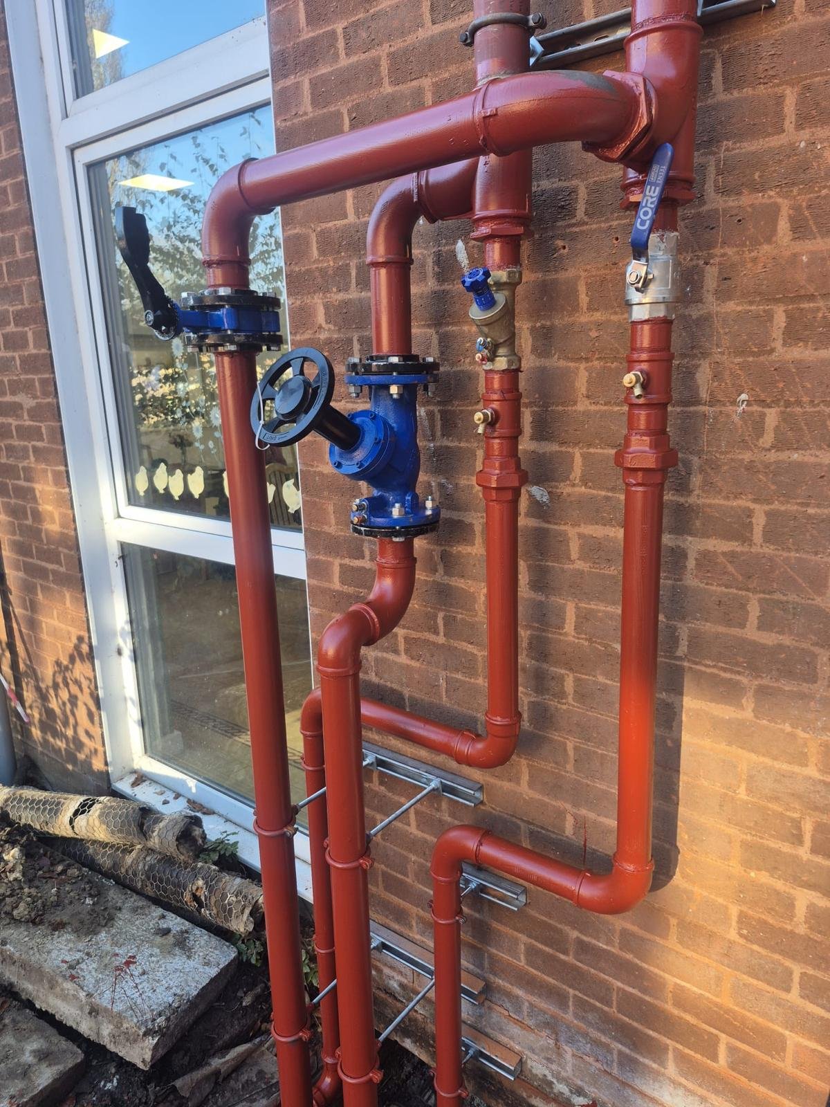 Red and blue plumbing pipes on a brick wall outside a building with a window.