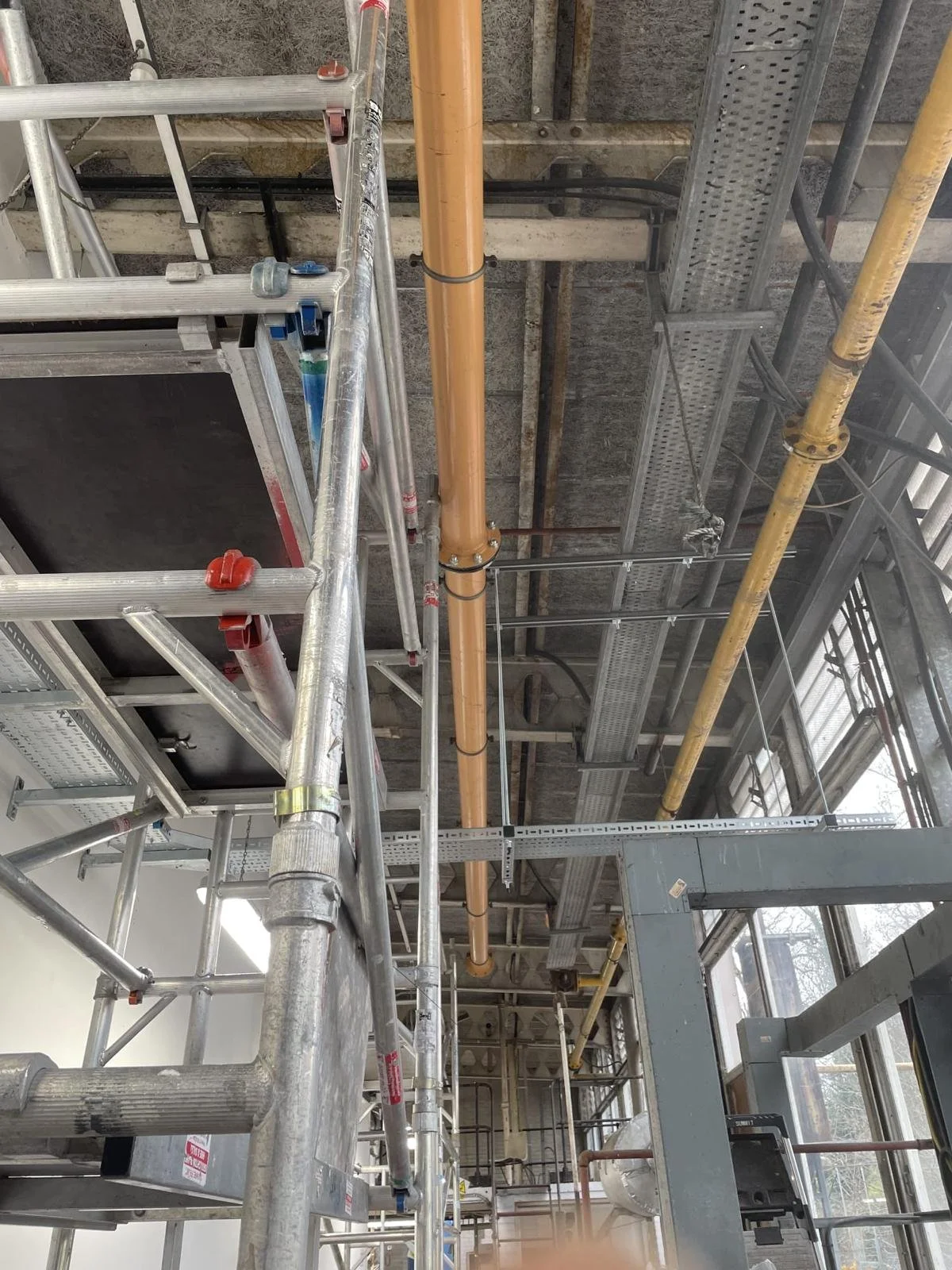 Interior view of a building under construction showing scaffolding, a beige plumbing pipe, yellow pipes, and metal framing.