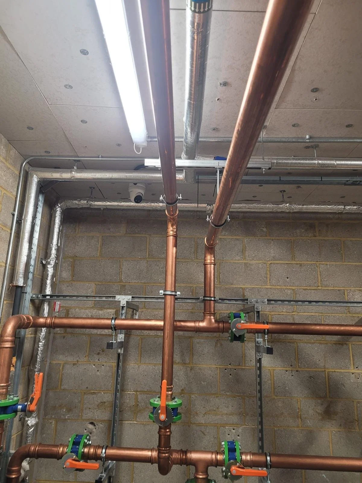 A plumbing installation with copper pipes and metal brackets mounted on a concrete wall, with a wooden ceiling and visible ceiling lights.