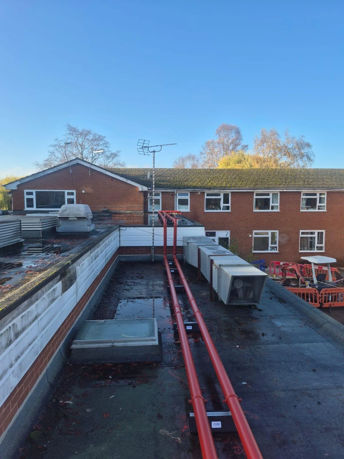View of a rooftop with red pipes, HVAC units, and a vent, overlooking neighboring brick buildings with windows, under a clear blue sky with some trees in the background.