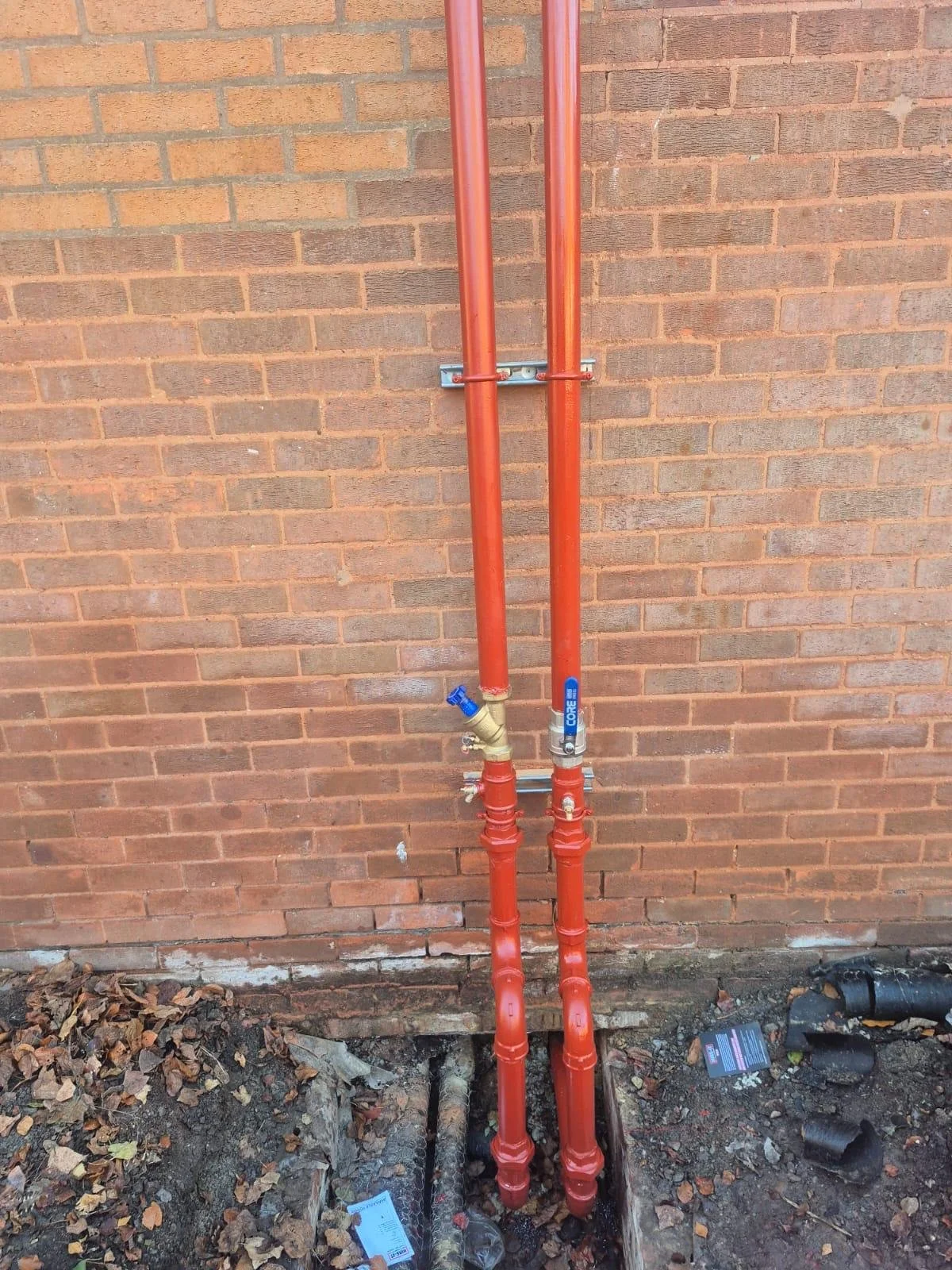 Red underground pipes with shutoff valves against a brick wall, surrounded by soil and leaves with some small debris.
