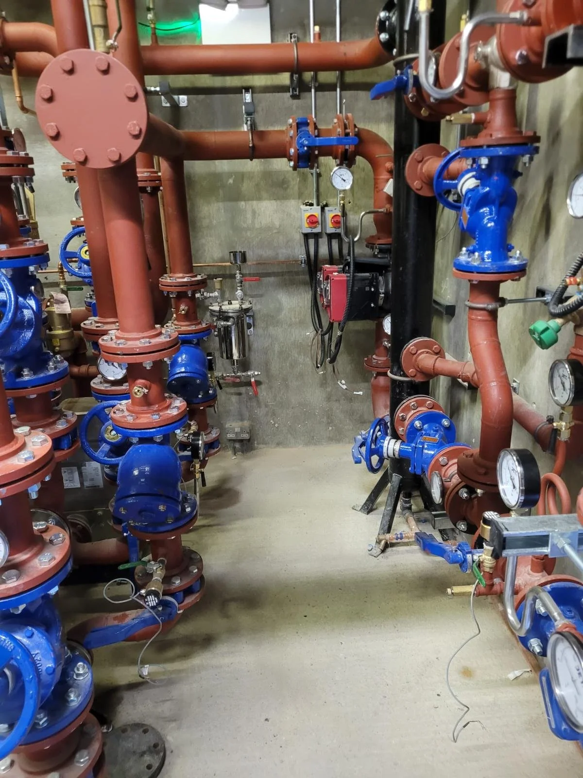 Industrial piping system with red and blue pipes, gauges, valves, and control devices mounted on a concrete wall and floor.