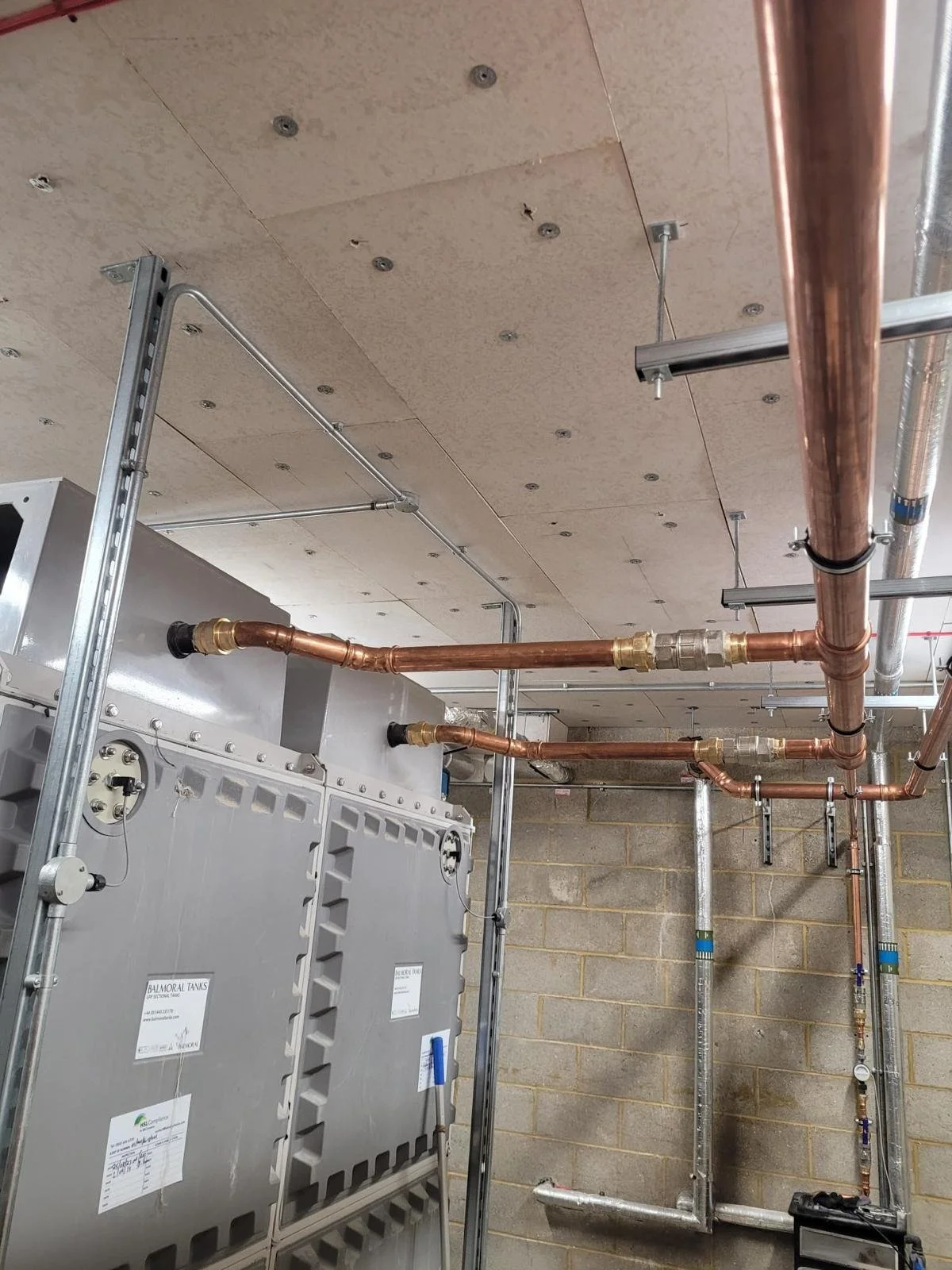 Construction site with copper pipes installed on a ceiling, connected to a large grey industrial unit, with metal scaffolding and a concrete wall in the background.