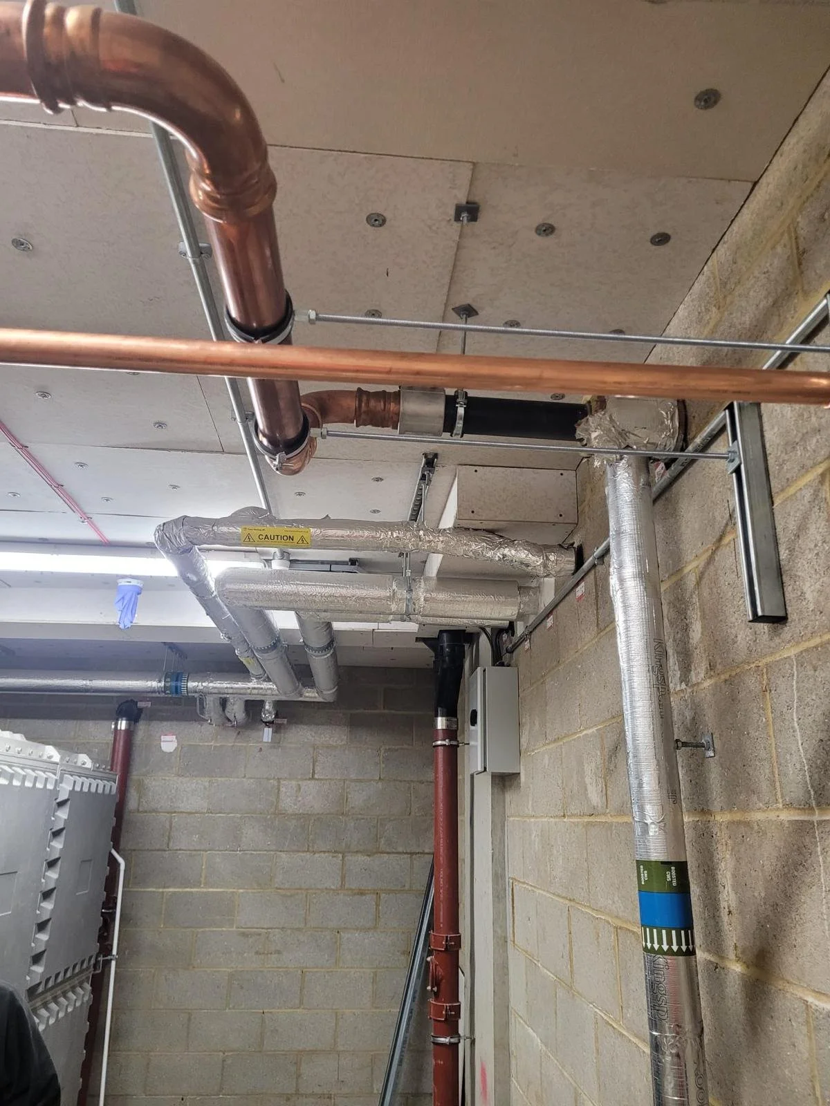 Construction area showing copper and insulated pipes, metal supports, and ductwork attached to a ceiling and walls.