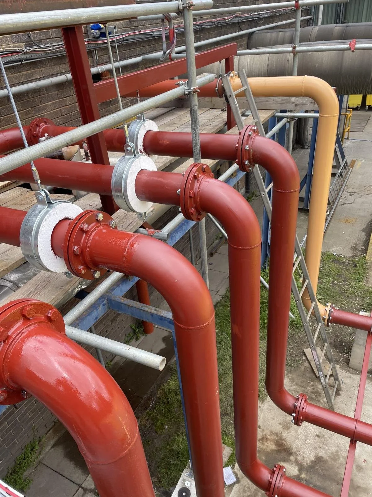 Red and beige pipes connected with flanges, supported by metal scaffolding at a construction site.