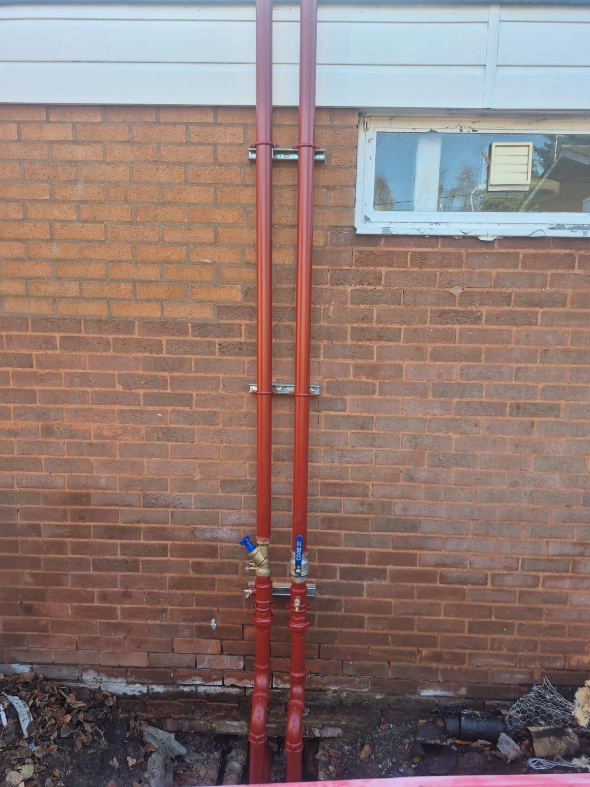 Red plumbing pipes run vertically along a brick wall next to a small window with a white frame.