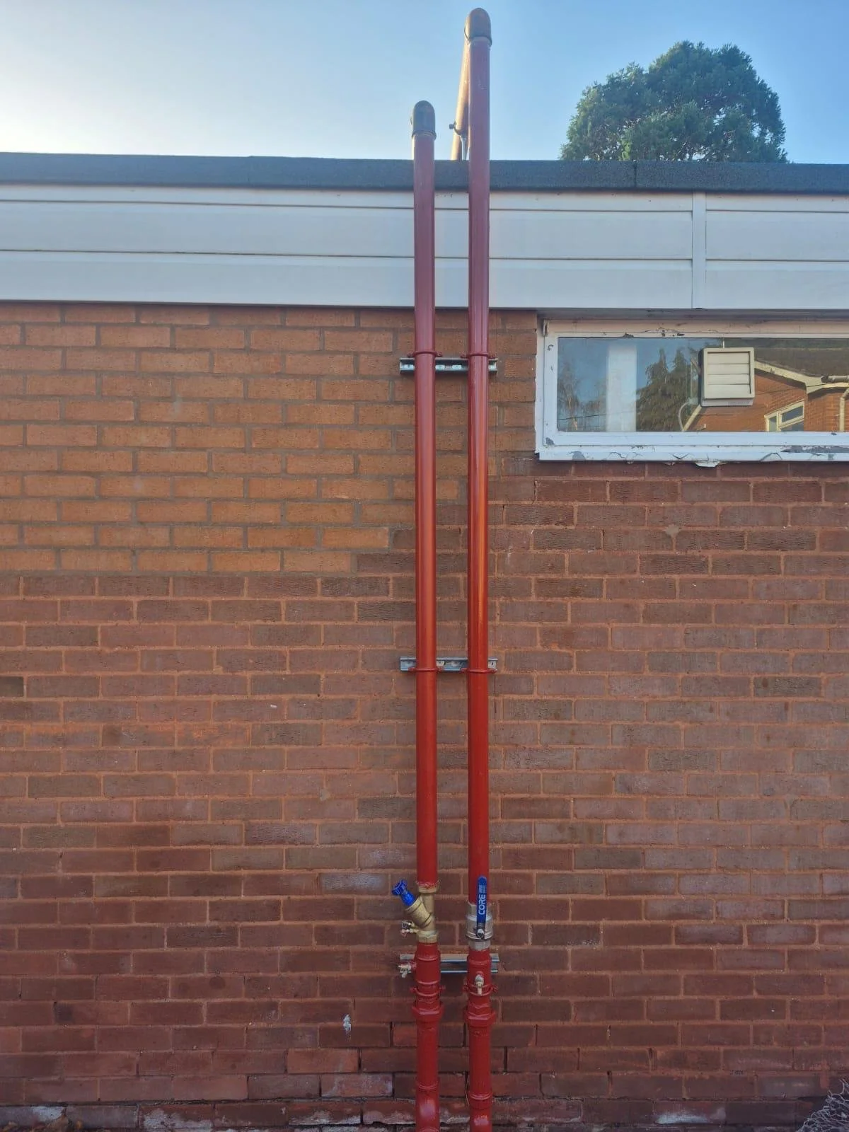 Two red industrial pipes mounted vertically on a brick wall beside a window with a tree in the background.