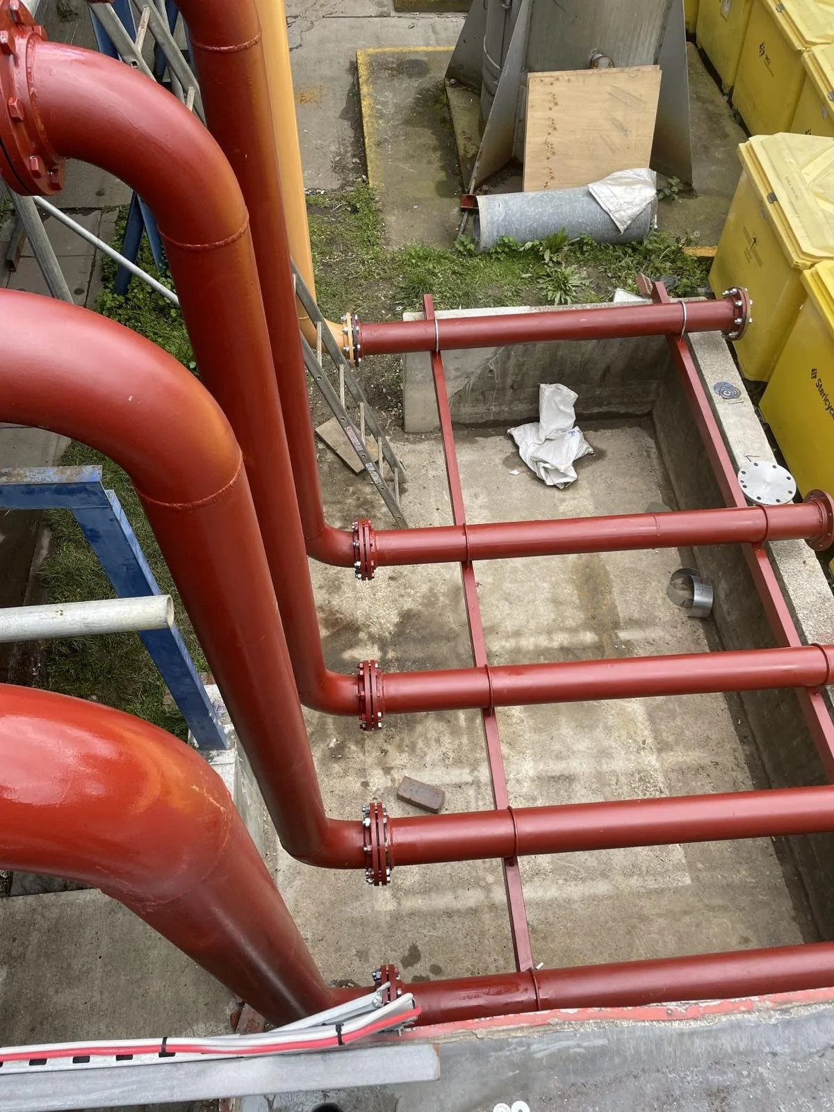 Red industrial pipes installed on a construction site with flanged fittings, over a concrete foundation, with a partially dug area and construction materials in the background.