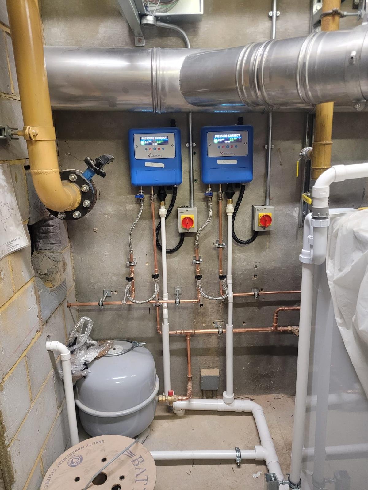 A plumbing and heating system with pipes, control units, valves, and gauges mounted on a concrete wall in a utility room.