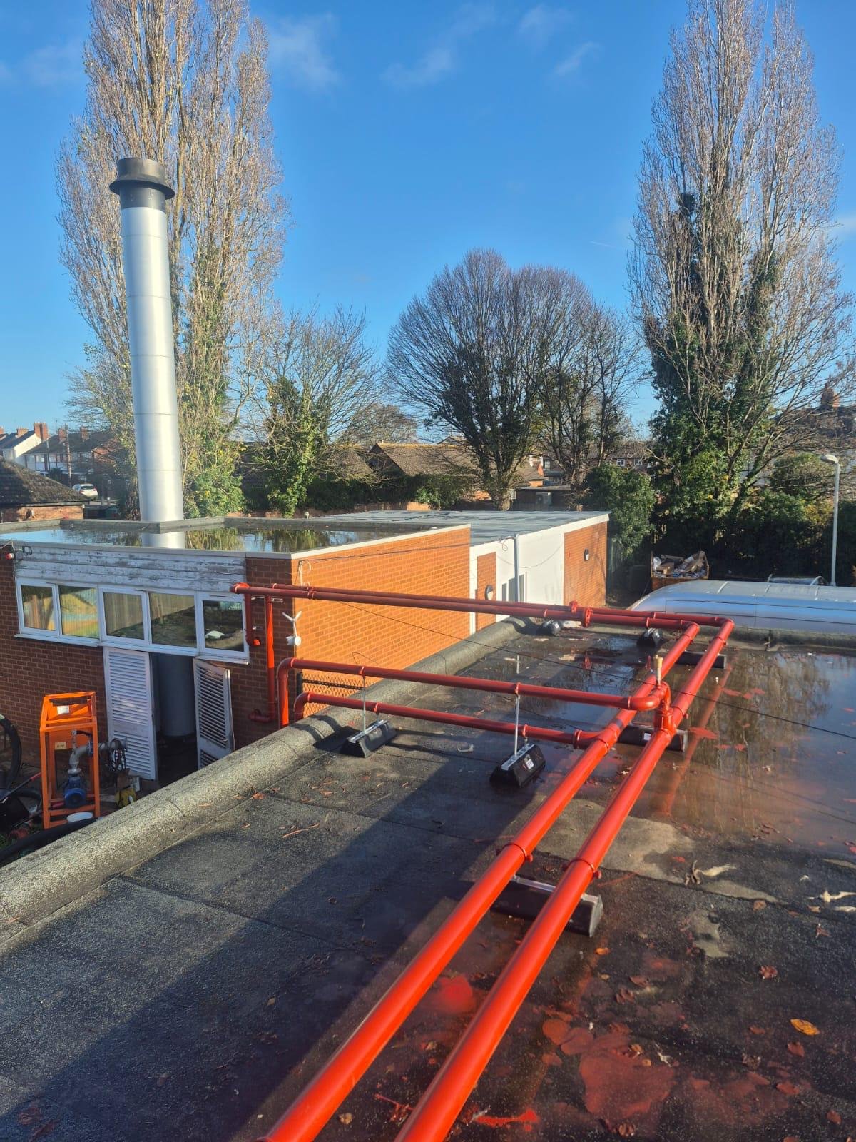 View of a rooftop with orange pipes, a reflective puddle, and nearby trees and buildings under a blue sky on a clear day.