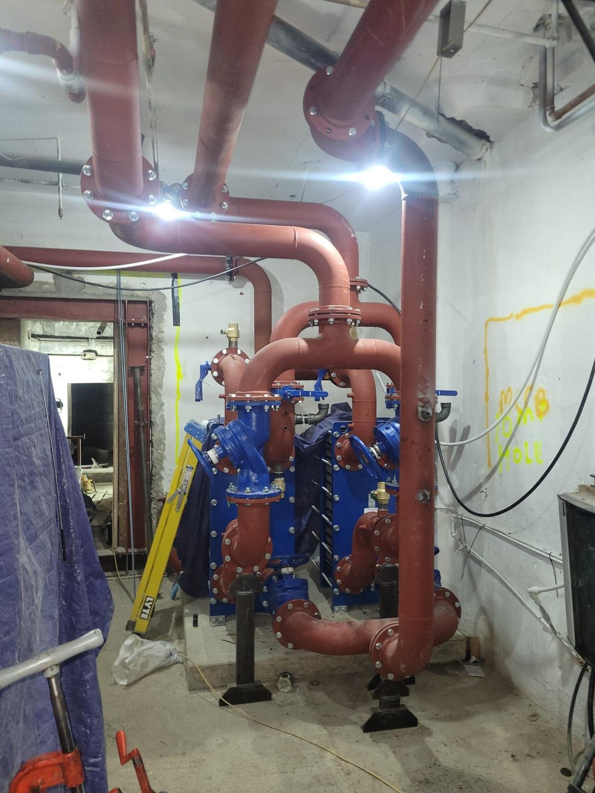 Construction site with orange and blue piping and valves, temporary support structures, and construction equipment.