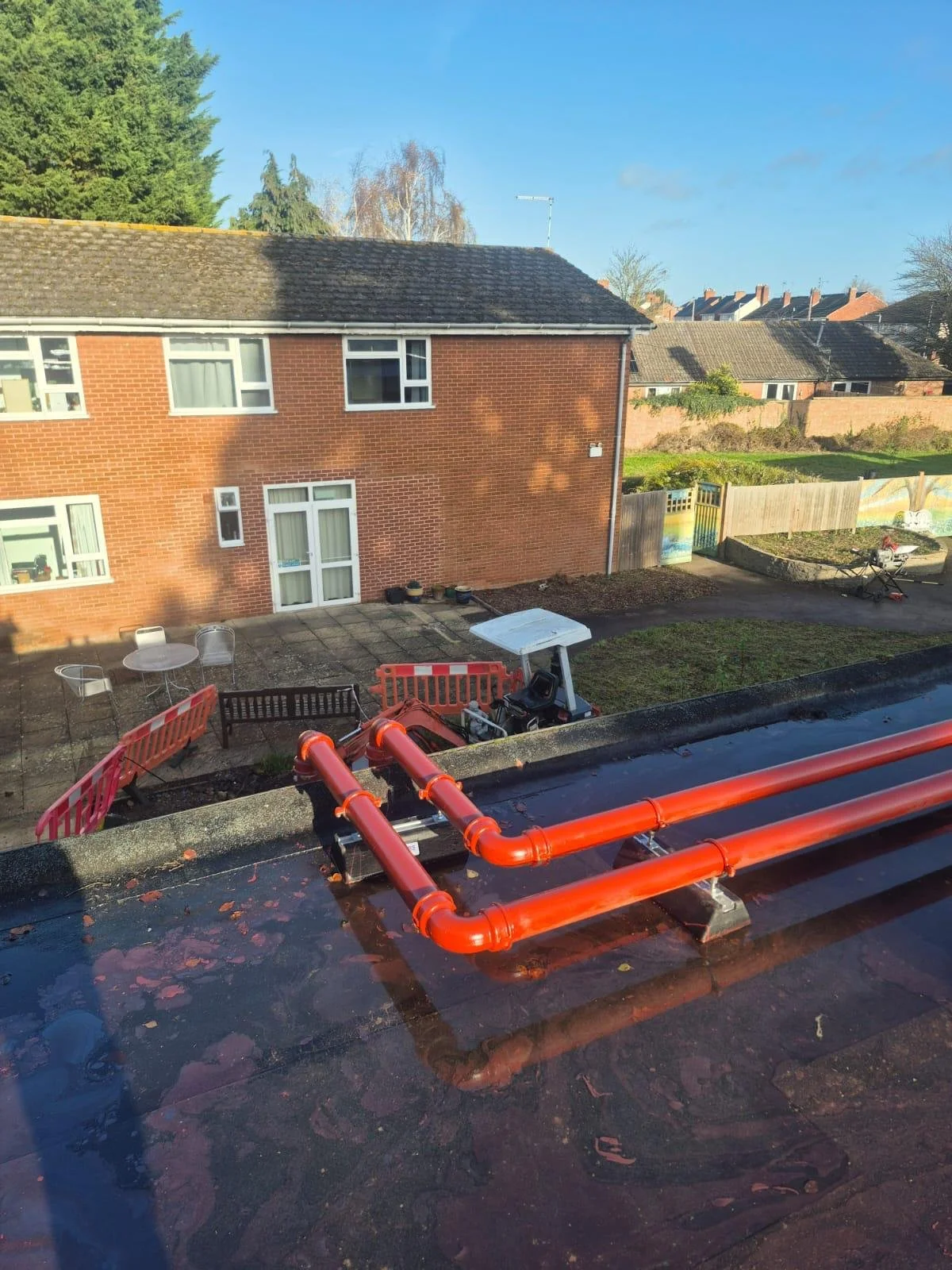 View from a window of a residential building showing a backyard with red pipes running across the roof, outdoor furniture, and a paved area. In the background, a brick house with a tile roof and other houses can be seen.