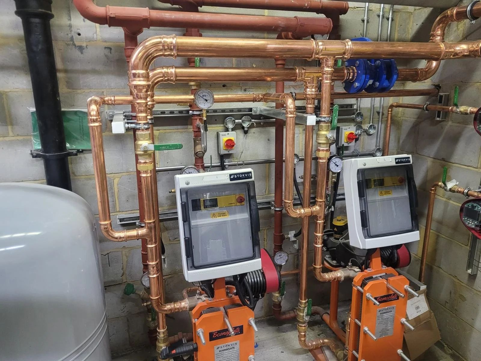 A mechanical room with copper piping, pressure gauges, and two white control panels mounted on orange devices.