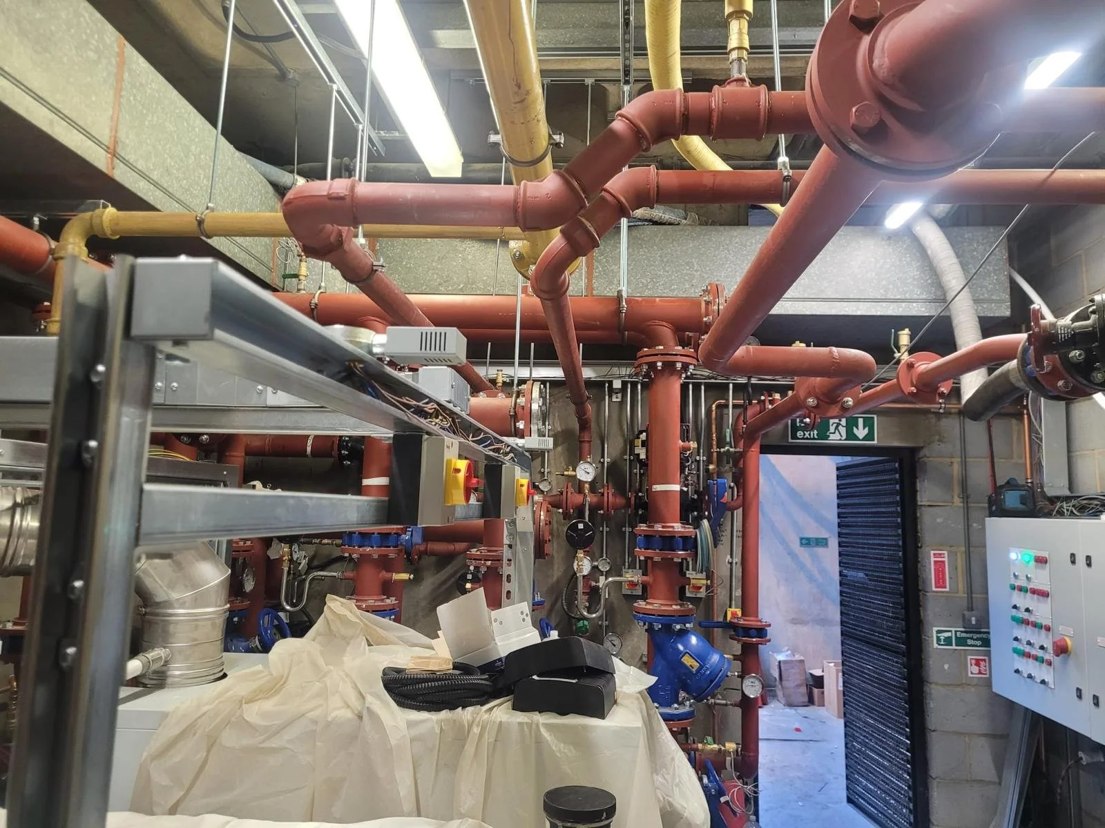 Photo of a complex network of red and yellow pipes and pressure gauges inside an industrial facility, with a partially open door and an emergency exit sign in the background.