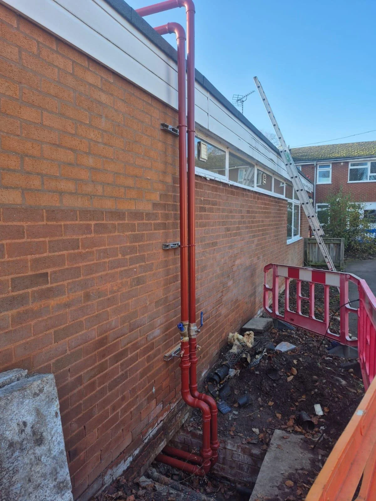 View of a building's exterior with visible red plumbing pipes, a ladder, a construction barrier, and an open trench below.