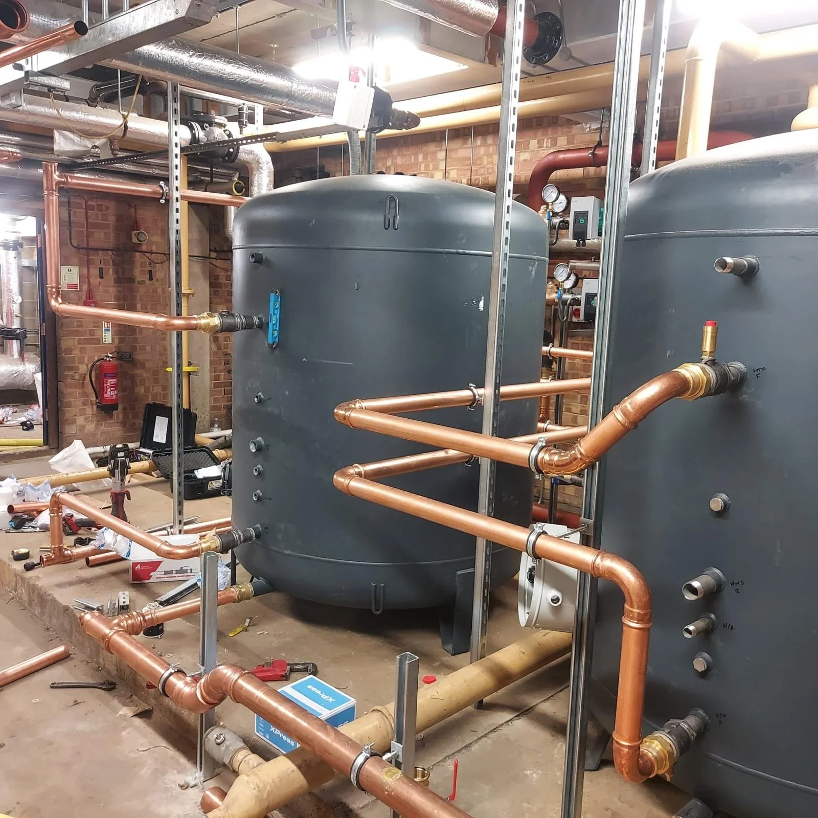 A heating, ventilation, and air conditioning system installation featuring multiple copper pipes, large gray tanks, gauges, and other HVAC equipment inside a building with brick walls.
