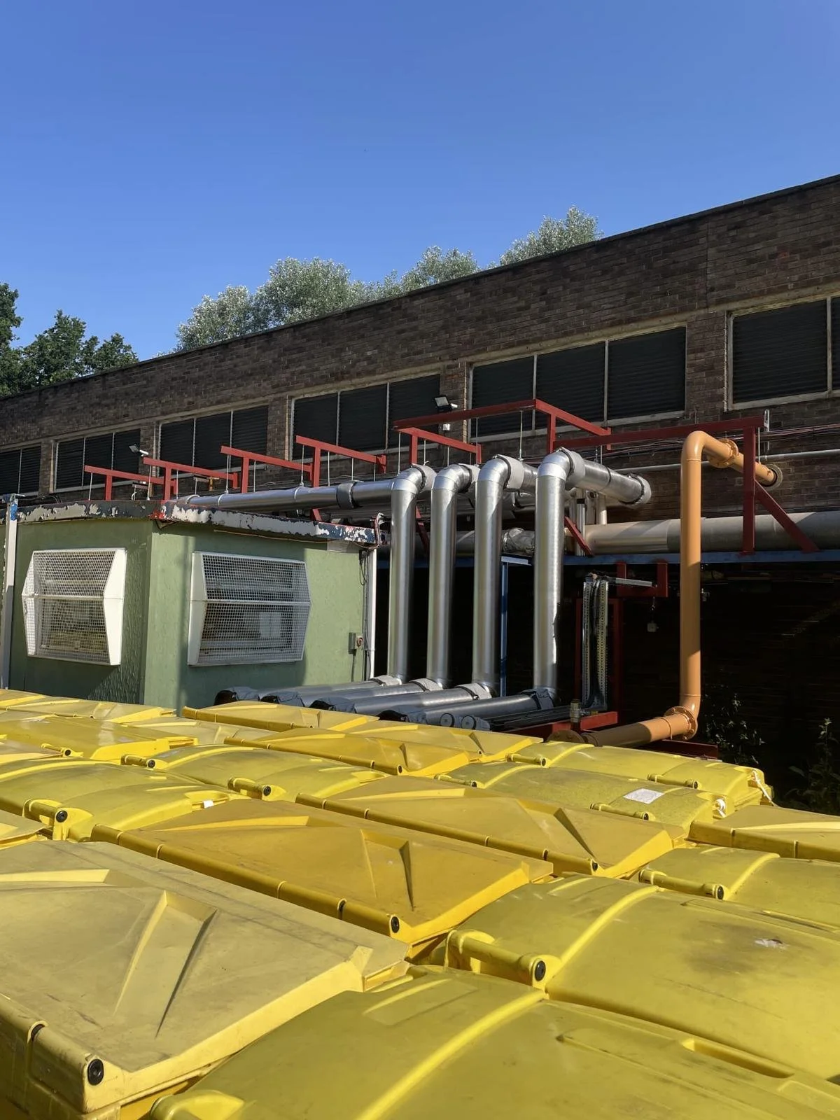 An industrial building exterior with metal pipes, vents, and yellow machinery or containers in front, under a clear blue sky.
