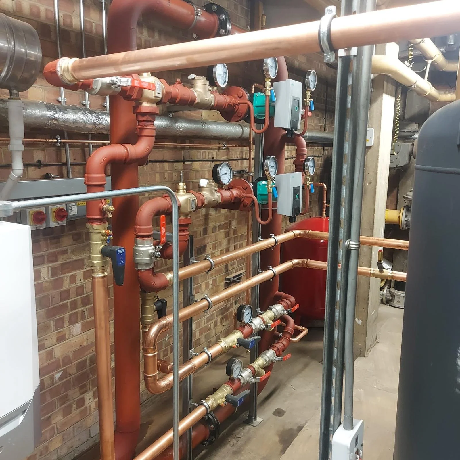 A mechanical room with various pipes, gauges, and valves, including copper and steel piping, mounted against a brick wall, with some electronic controllers and gauges.