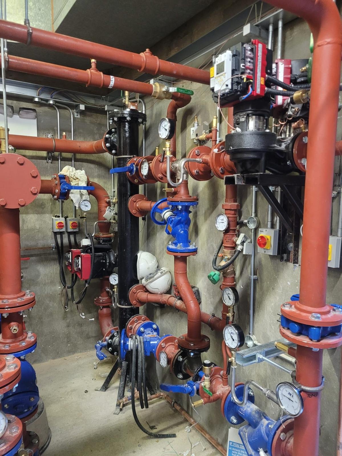 A complex system of plumbing pipes, gauges, and control valves in an industrial setting, with various valves, meters, and electronic controls mounted on a concrete wall.