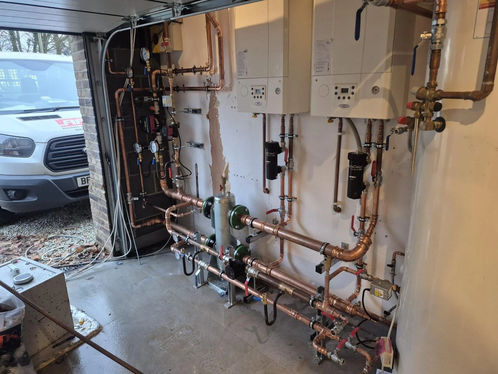 View of a heating or plumbing system installed indoors, with copper pipes, gauges, valves, and control units. A partially open garage door revealing an outside vehicle.