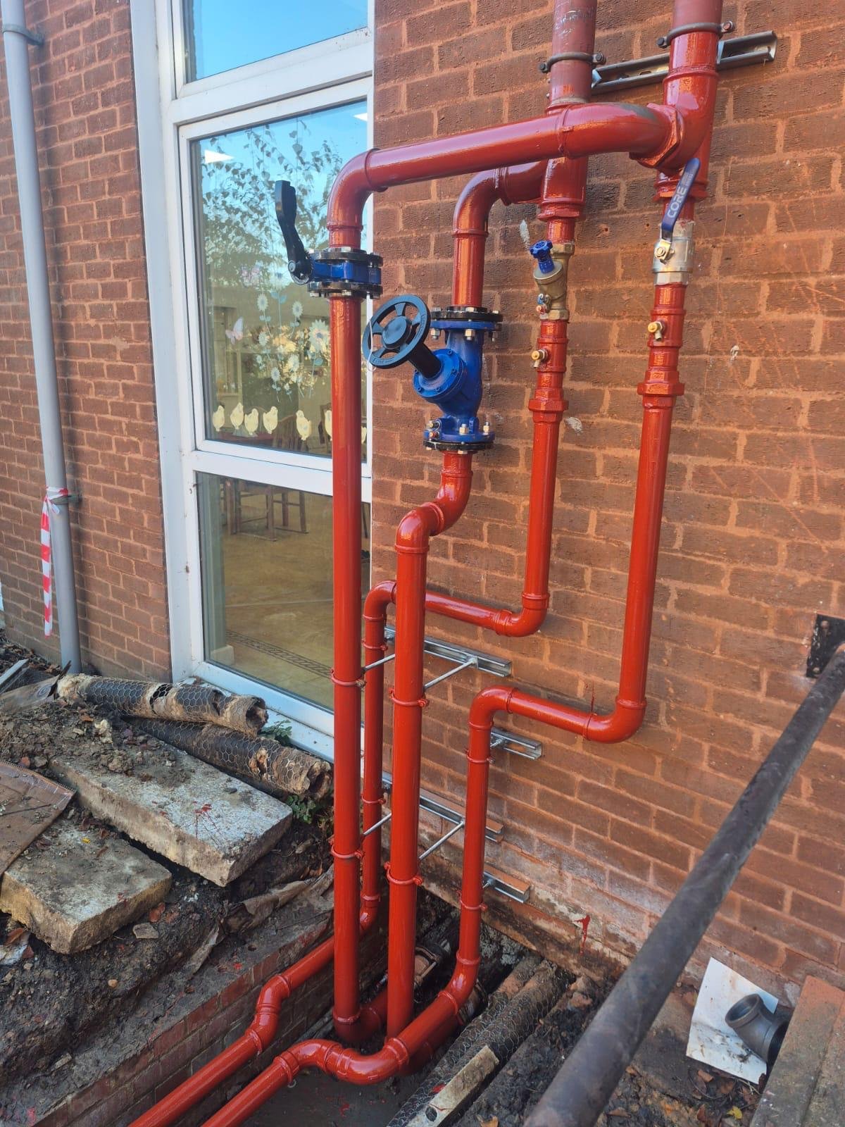 Red piping system with valves installed outside a brick building, next to a window, with some construction material and dirt on the ground.