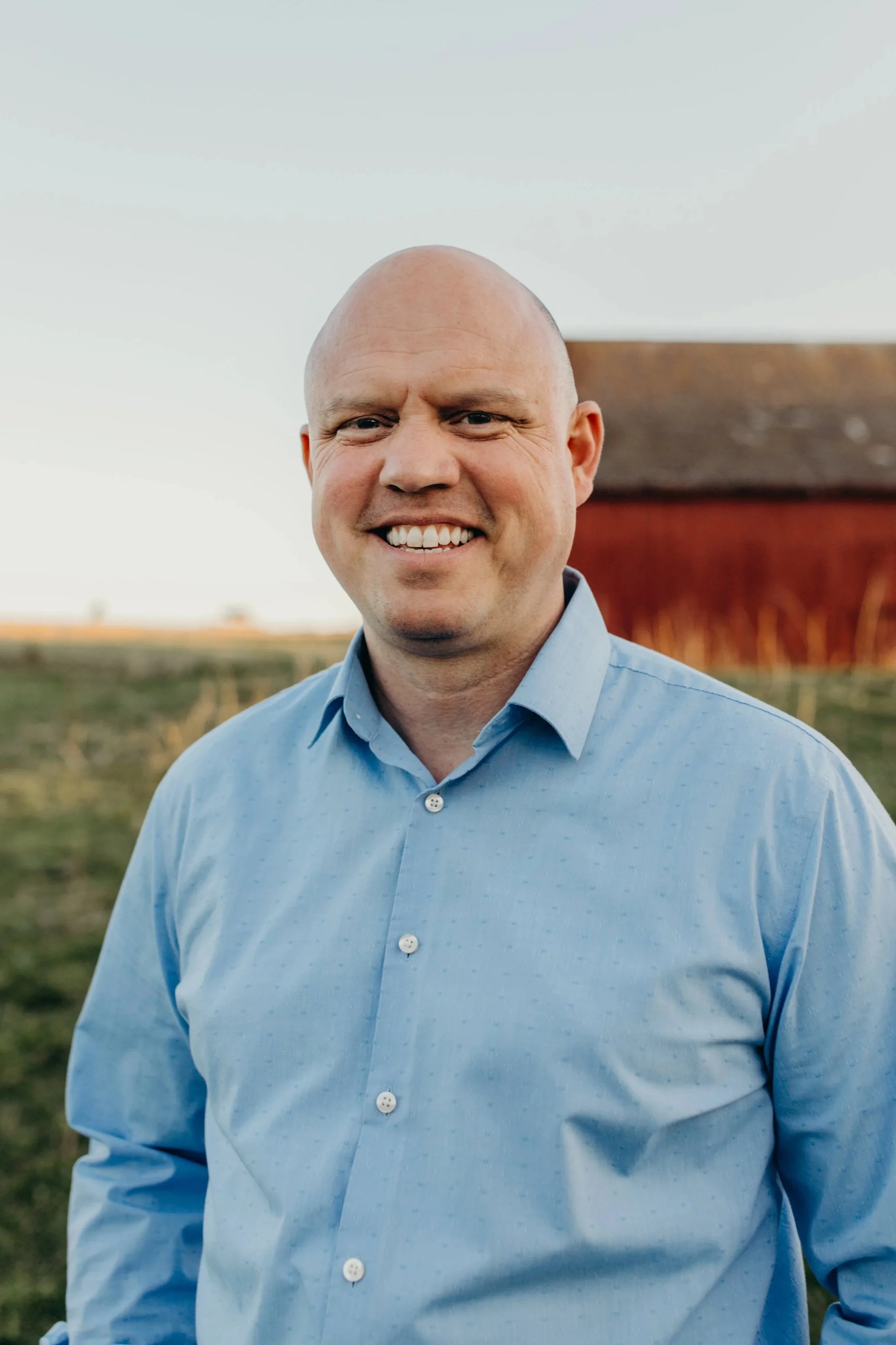 A smiling man standing outside in front of a wooden structure, wearing a light blue button-up shirt.