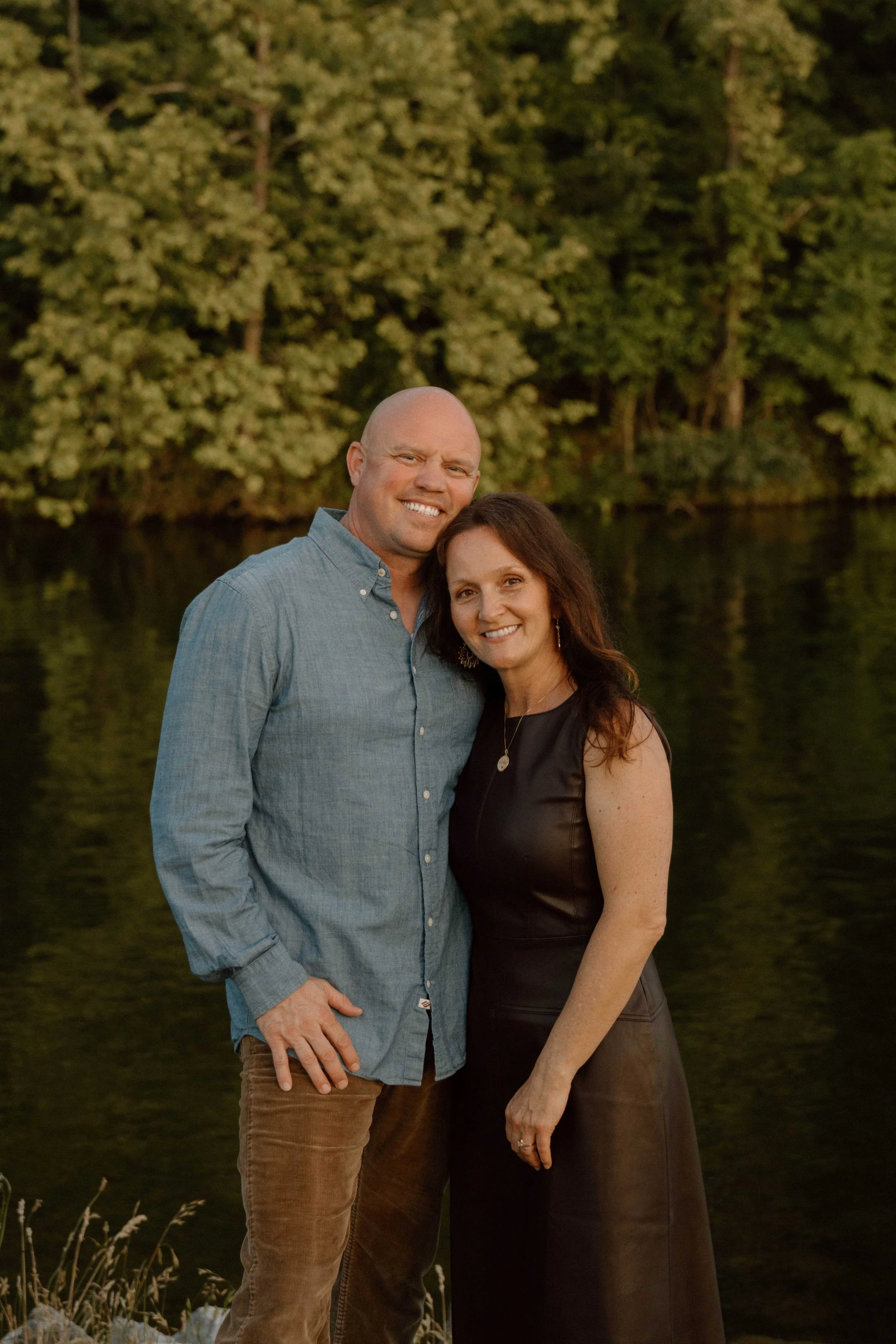 A smiling man and woman standing outdoors near water, with green trees in the background during sunset.