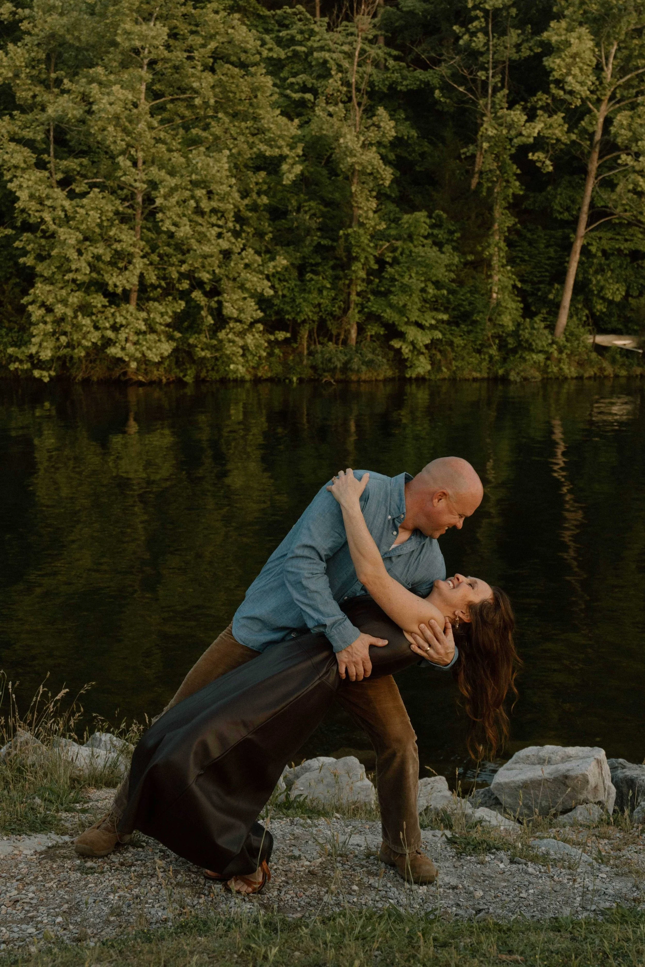A man and woman are performing a dance pose near a body of water, with the man leaning forward and the woman leaning backward, holding each other. They are standing on a rocky shoreline with grass and stones, surrounded by trees and reflecting water.