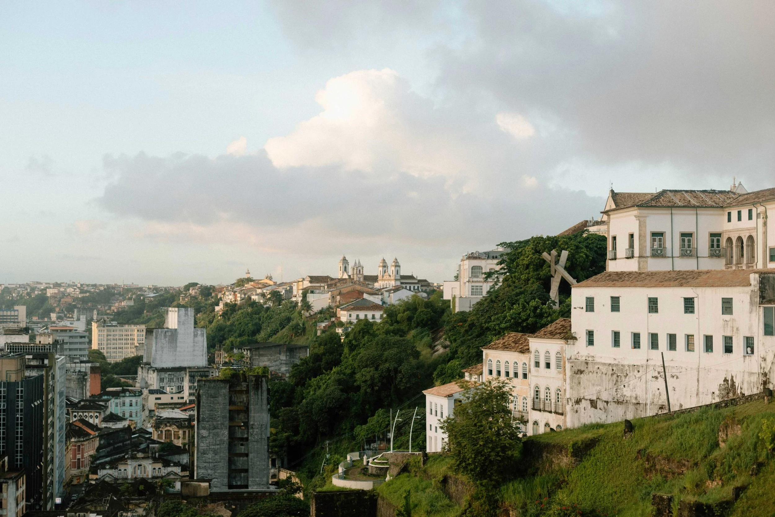 View of a cityscape with historic buildings, including churches with twin towers, surrounded by greenery and modern structures, under a cloudy sky.