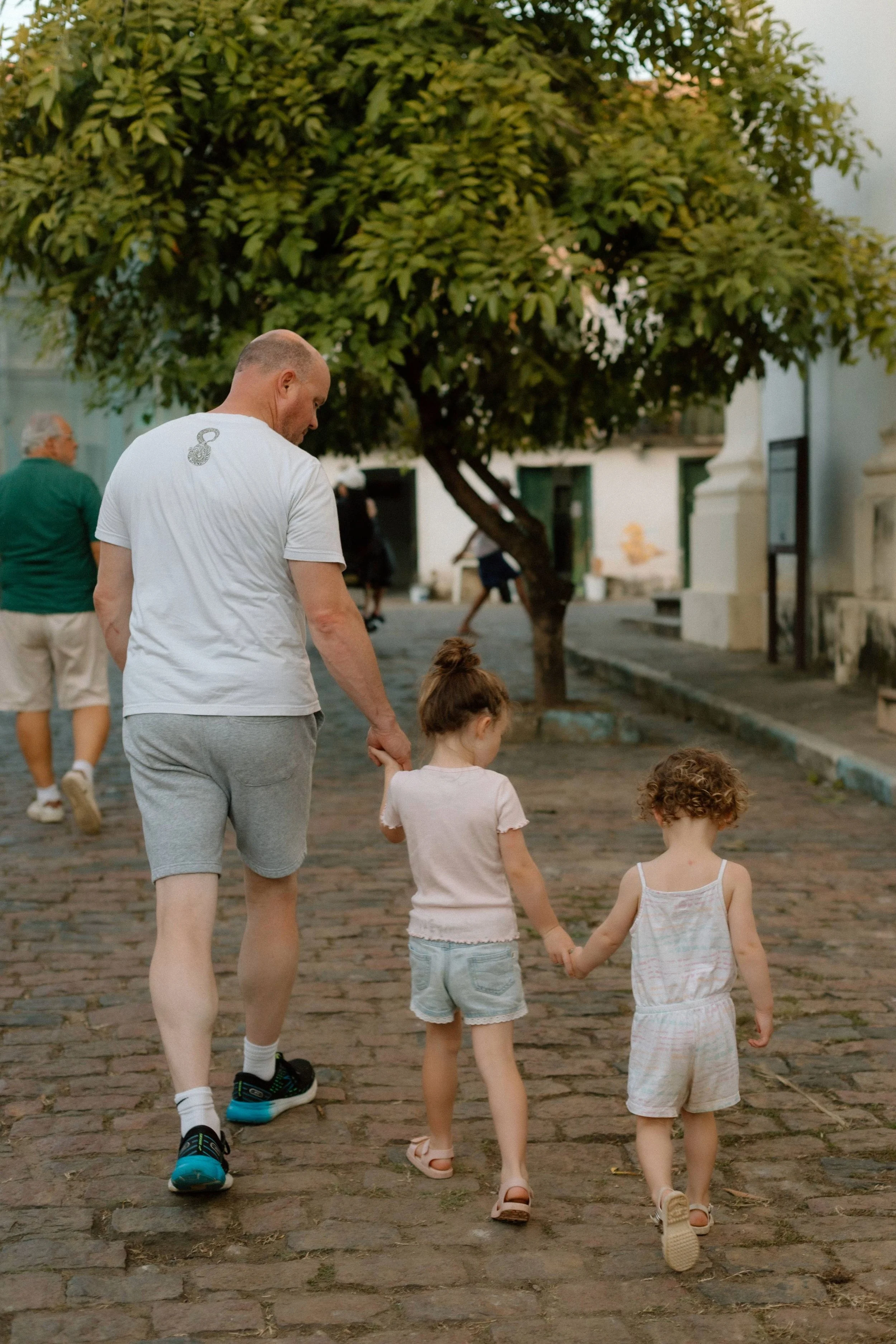 A man walking with three young girls holding hands on a cobblestone street, with a tree and some buildings in the background.