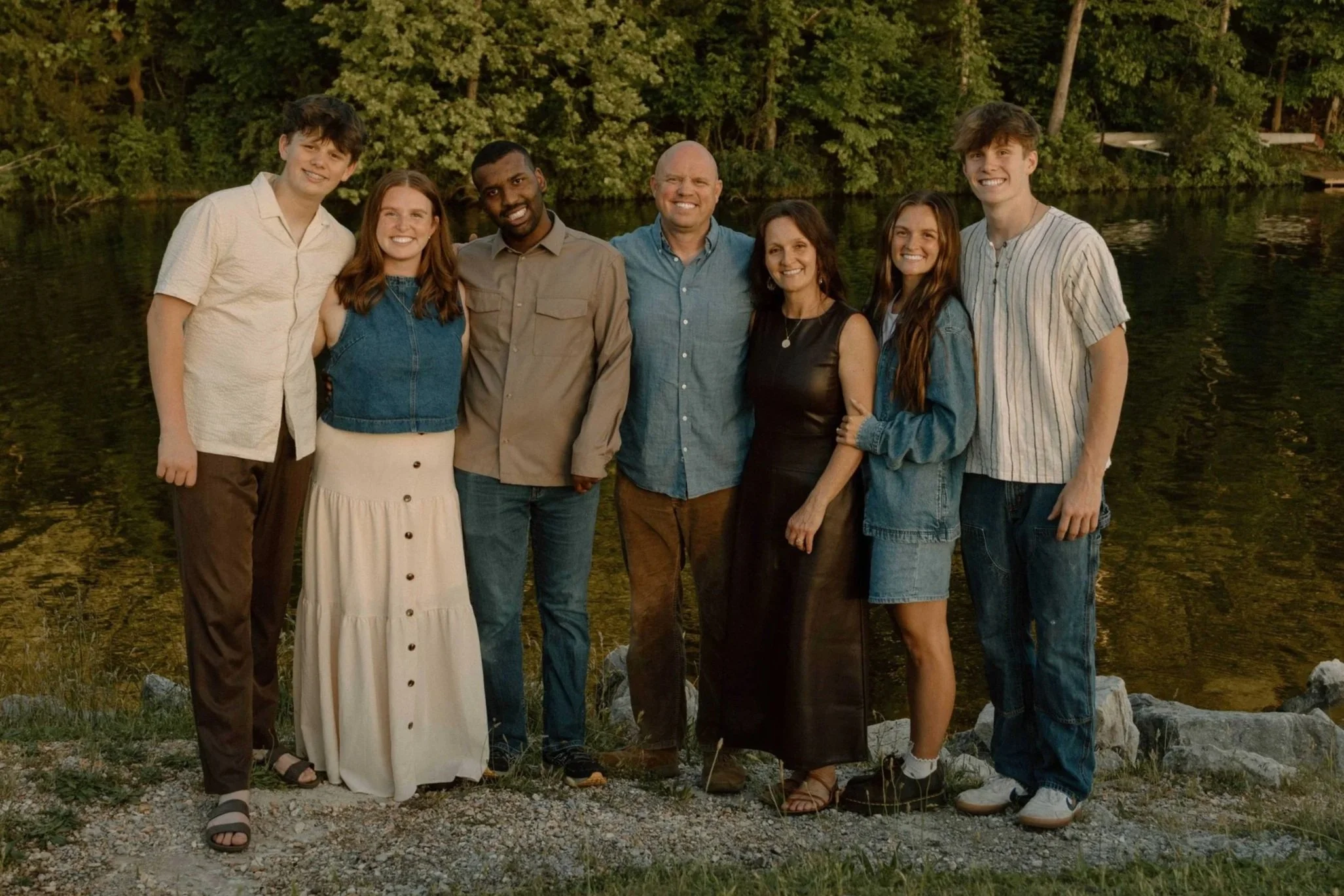 Group of seven people standing outdoors near a body of water with trees in the background, smiling and posing for a photo.