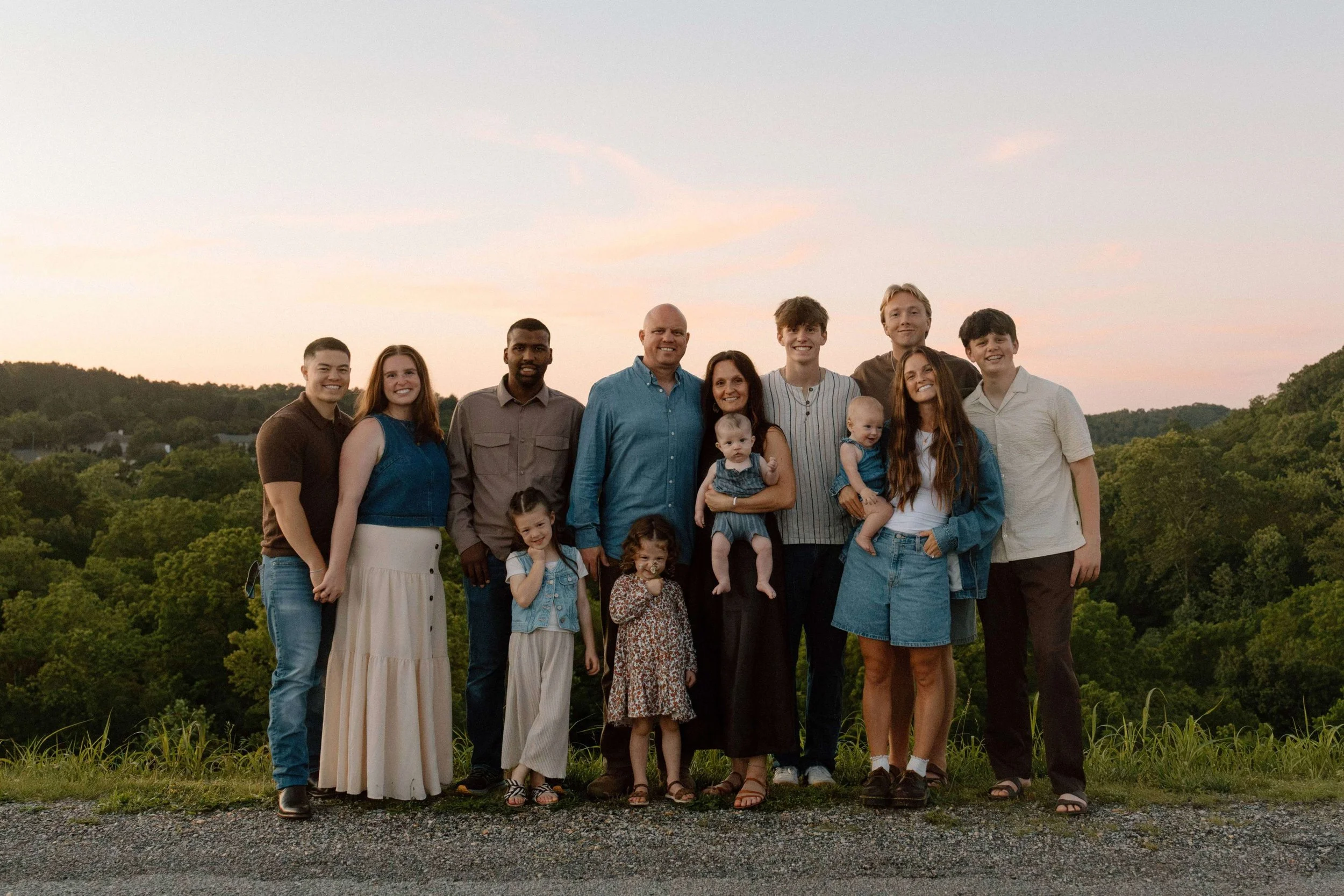 A diverse group of ten people, including children and adults, standing outdoors on a grassy area during sunset, with trees and hills in the background, smiling and posing for the photo.