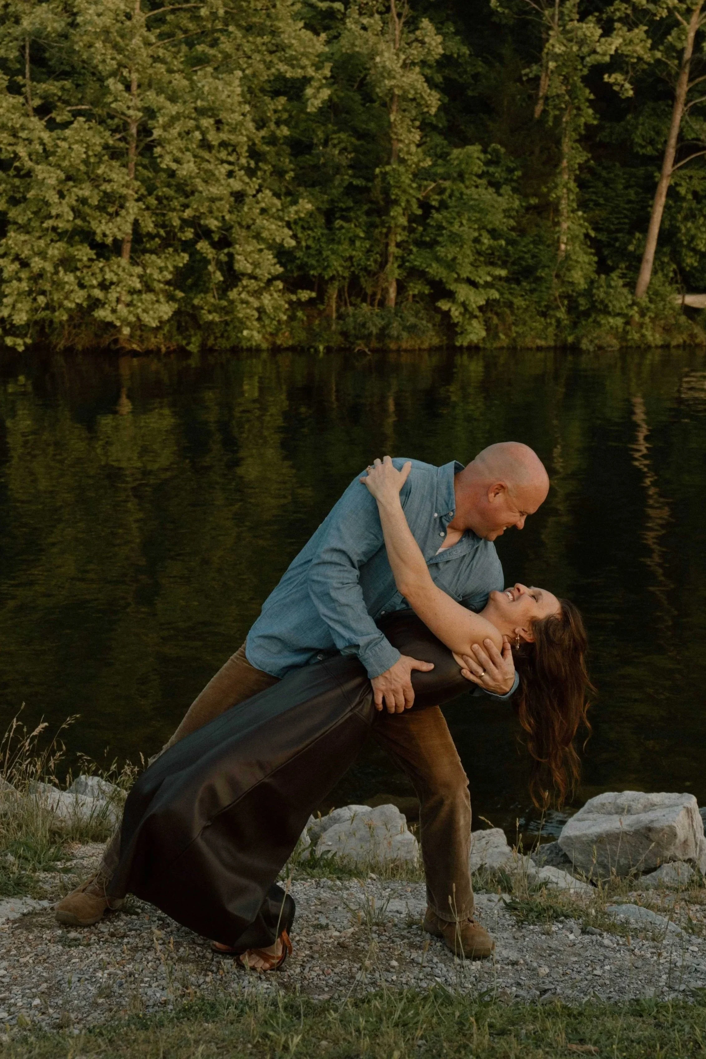 A couple is dancing outdoors near a body of water with a forest in the background. The man is bald, wearing a blue shirt, and the woman has long brown hair, wearing a black top and brown pants. They are engaged in a close, intimate dance, with the man dipping the woman back.
