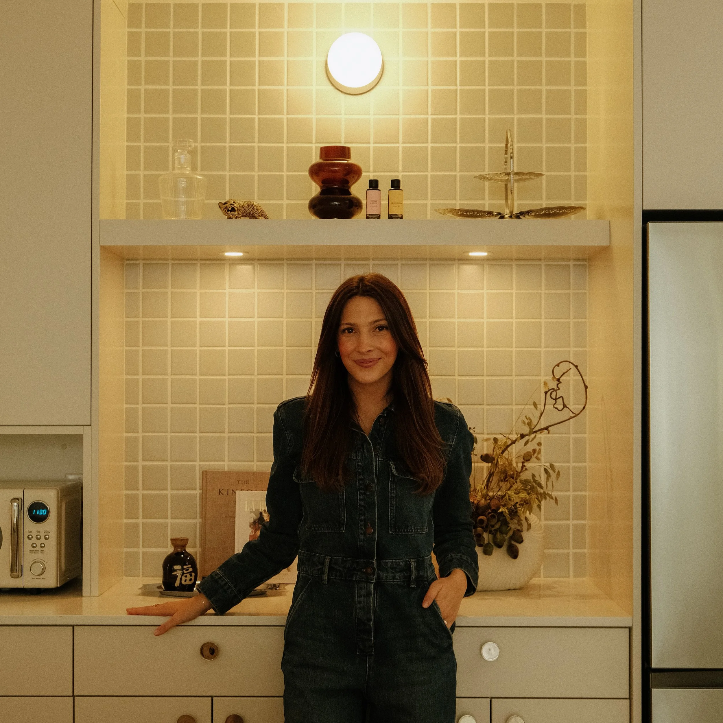 A woman with long dark hair and a denim jacket standing in a kitchen, smiling at the camera. Behind her are beige tiled walls with decorative items on a shelf, including a ceramic vase, a small leopard figurine, bottles, and a two-tiered tray. Part of a microwave, a clock, and a dried flower arrangement are also visible.