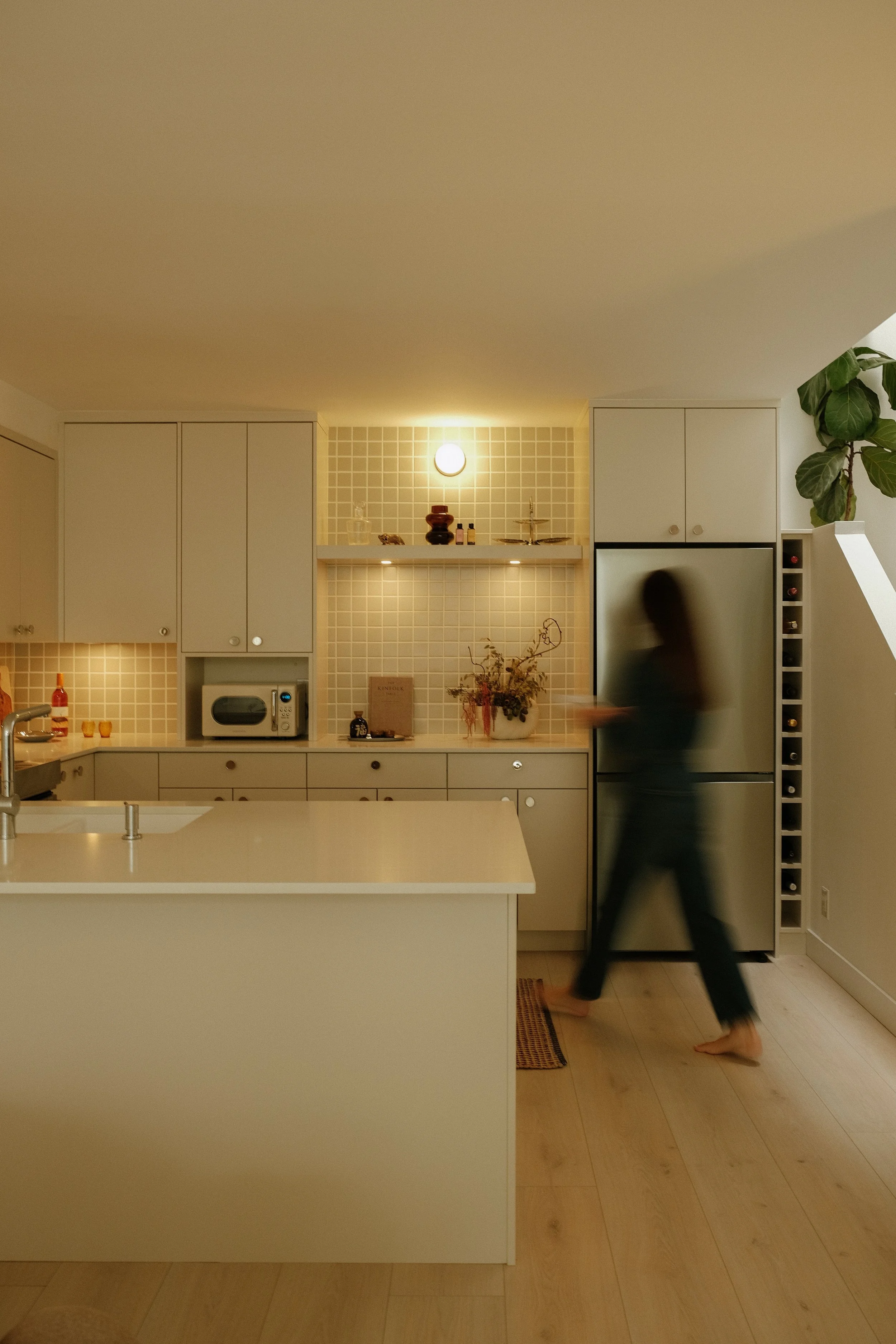 A modern kitchen with white cabinets, beige tiled backsplash, and a kitchen island. A person with dark hair dressed in black is walking barefoot, slightly blurred, past a stainless steel refrigerator. Decor items including plants, bottles, and a microwave are visible.
