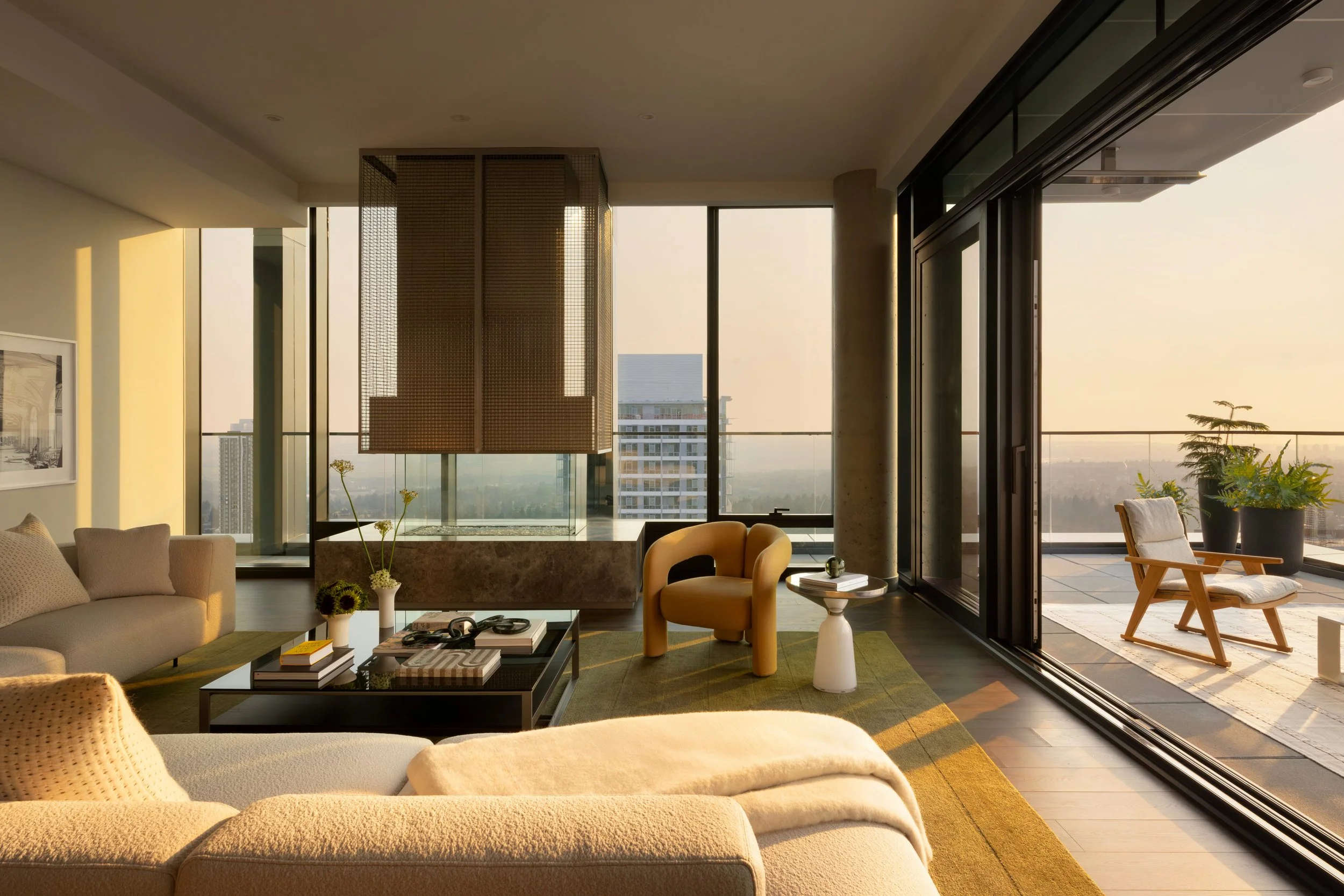 Modern living room with beige sofas, black coffee table, yellow armchair, white side table, large floor-to-ceiling windows with a city view, balcony with chairs and plants, and warm sunlight.