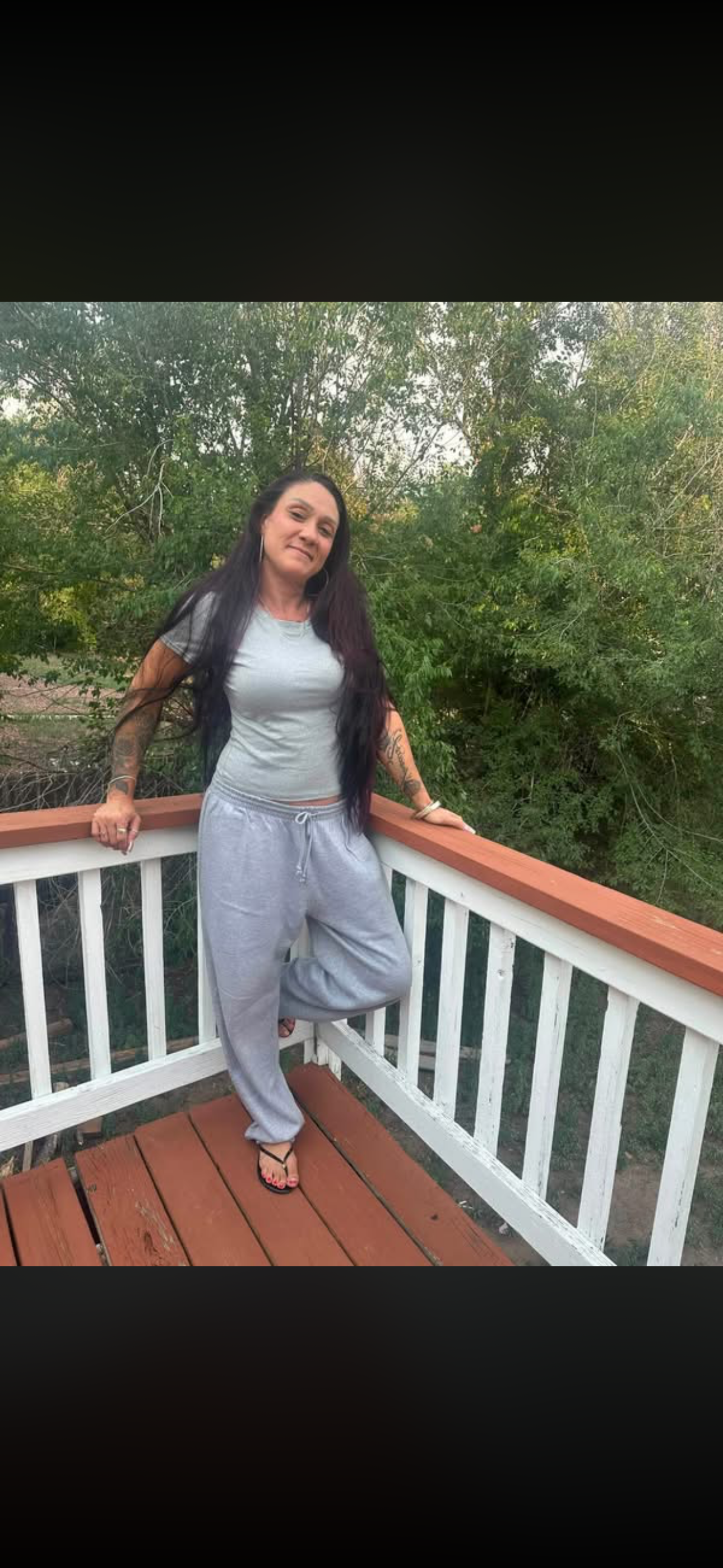 Woman with long dark hair standing on a wooden deck, leaning on the railing, outdoors with green trees in the background, wearing a gray t-shirt, gray sweatpants, and black flip-flops.