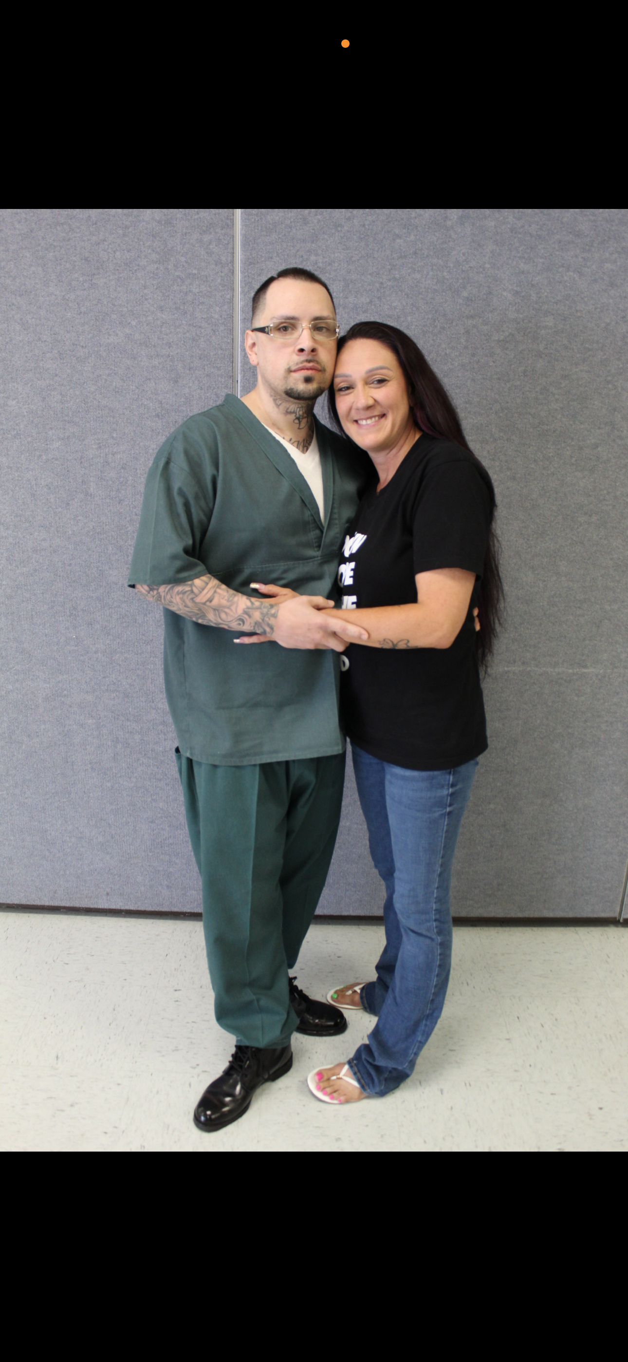 A man in green scrubs and a woman in casual clothing smiling and holding hands against a gray wall.