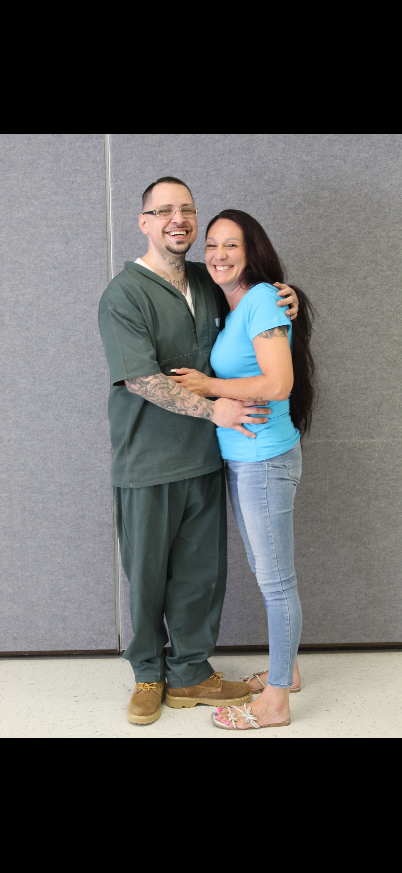 A man and woman smiling and embracing each other, standing against a plain gray wall.