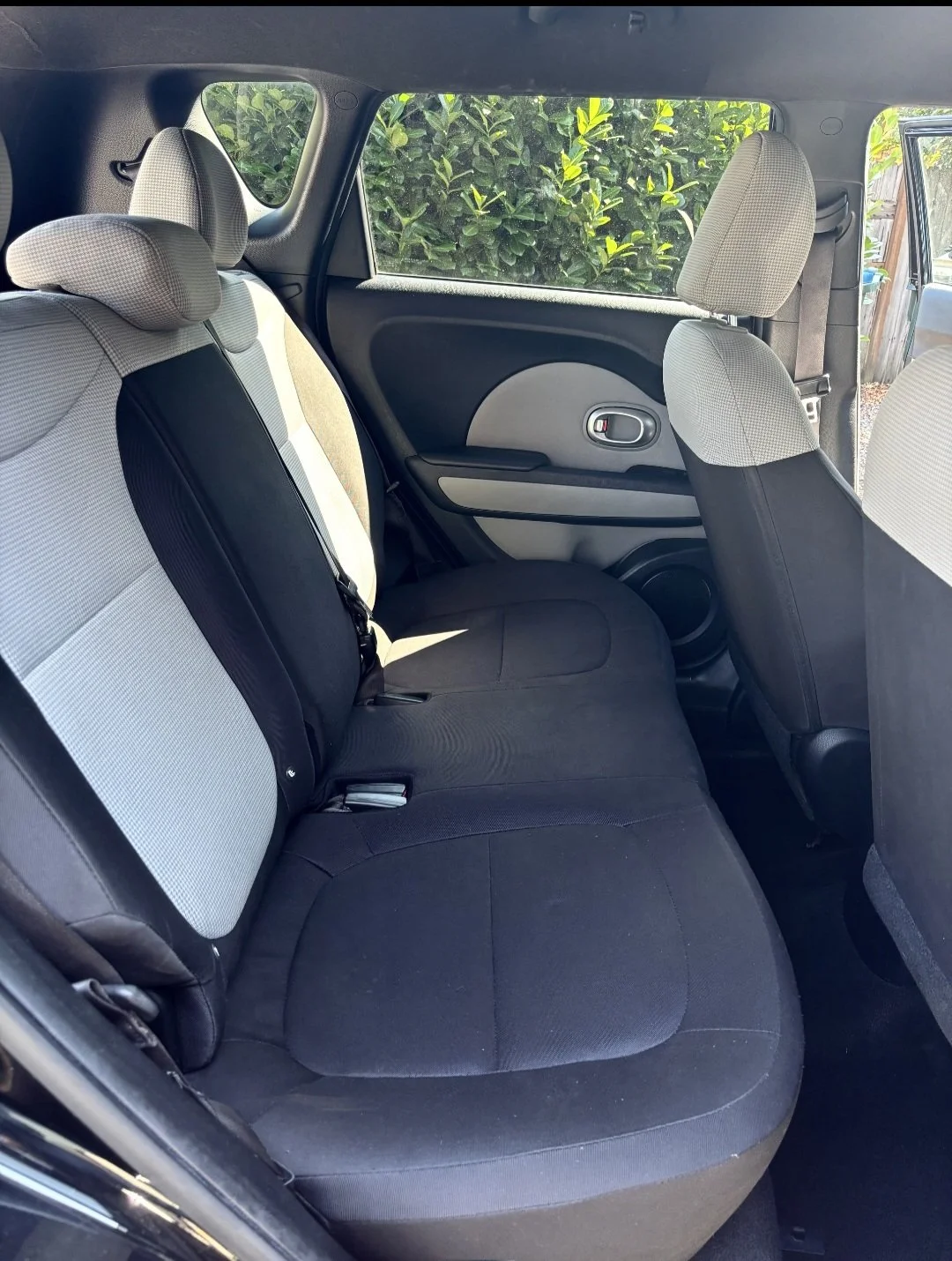 The back seat of a compact car showing black and beige upholstery, a window with greenery outside, and a partially visible front seat.
