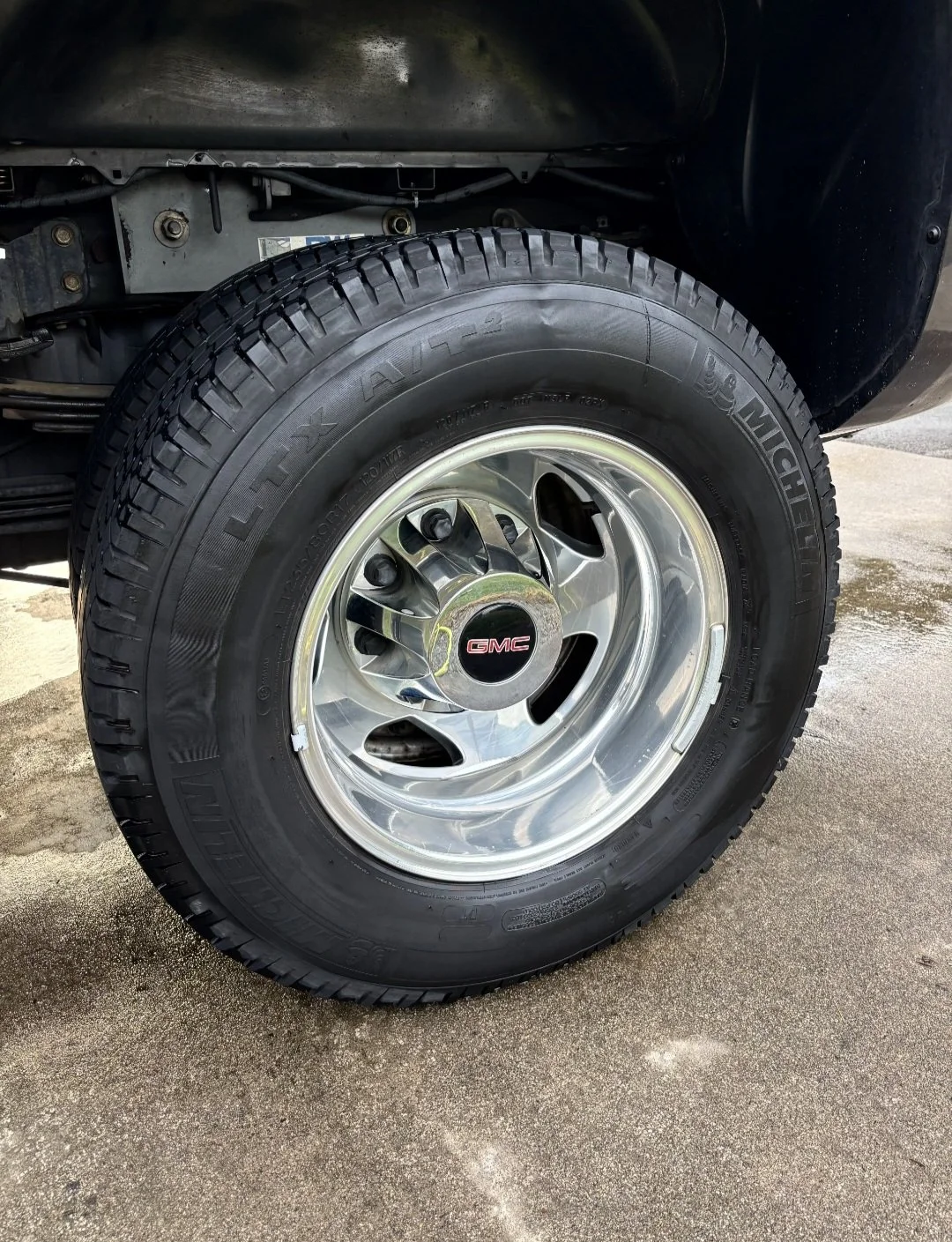 Close-up of a GMC truck wheel with a black tire and shiny chrome rim, mounted on the vehicle's front axle, with a visible GMC logo in the center cap.