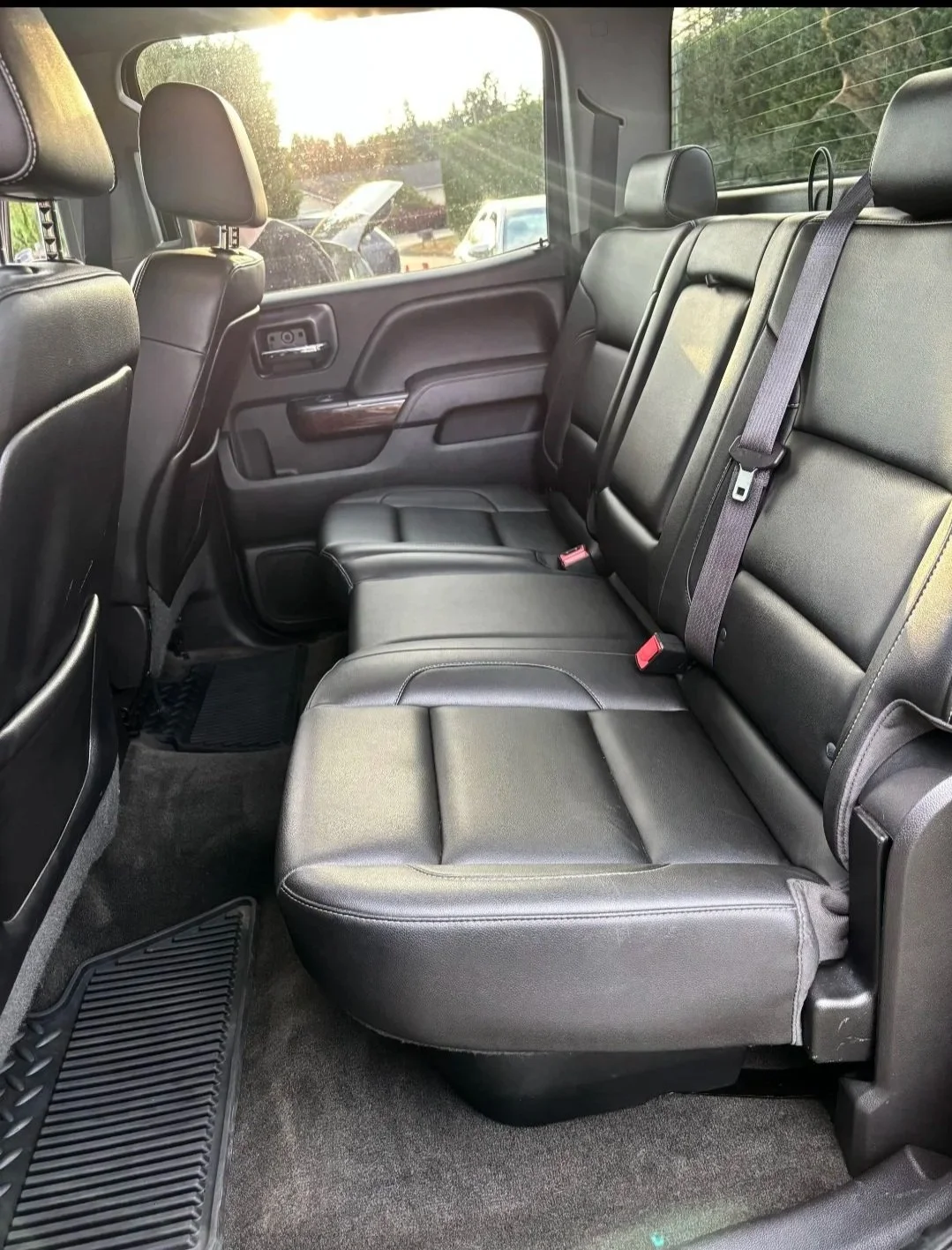 Interior view of the backseat of a vehicle with black leather seats, a seatbelt, and a rear window showing a sunny day outside with trees and cars.