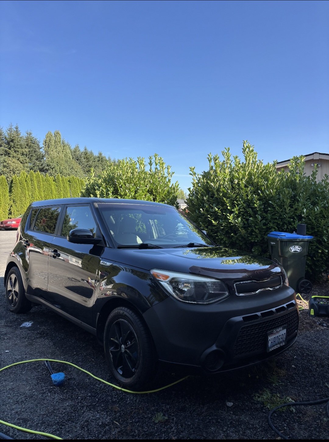 A black Kia Soul parked outdoors on a gravel driveway, with green bushes and trees in the background under a clear blue sky.