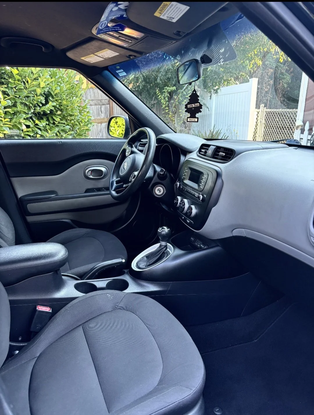 Interior of a car showing the driver's seat, steering wheel, dashboard, and center console with gear shift. The view is from the passenger side, with a lush green bush and a wooden fence visible outside the vehicle.