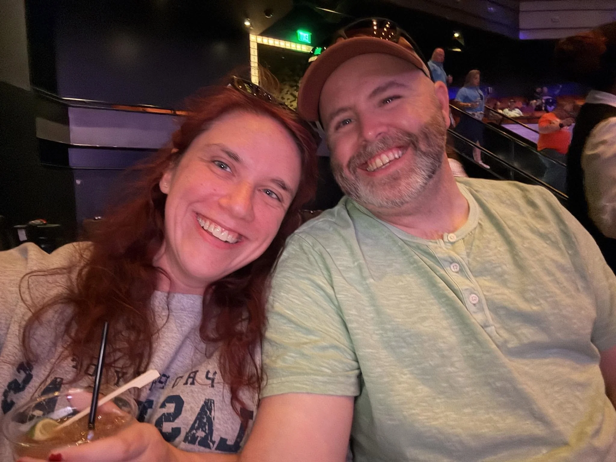 A woman with long red hair and a man with a beard and a tan cap taking a selfie in a dimly lit indoor amusement park or entertainment venue, with people and arcade game machines visible in the background.
