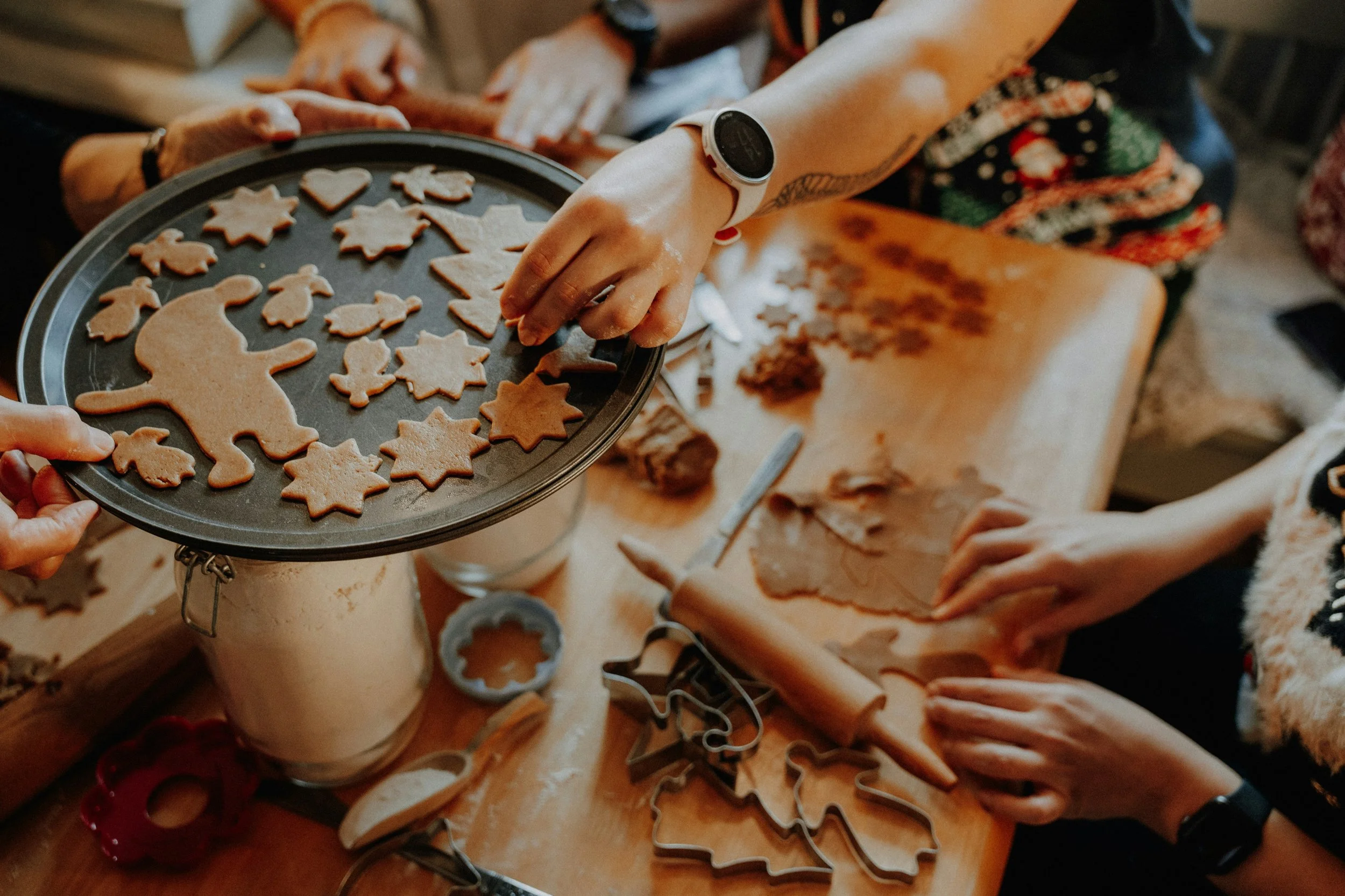 Multiple cookies in various holiday shapes such as stars, trees, and gingerbread men, with some cookies on a tray and others on a wooden surface. People are decorating or preparing the cookies in a festive setting.