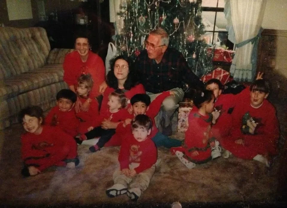 A family gathered for Christmas in front of a decorated Christmas tree with presents. The group includes adults and children wearing red Christmas sweaters, sitting on a carpeted floor inside a cozy home.