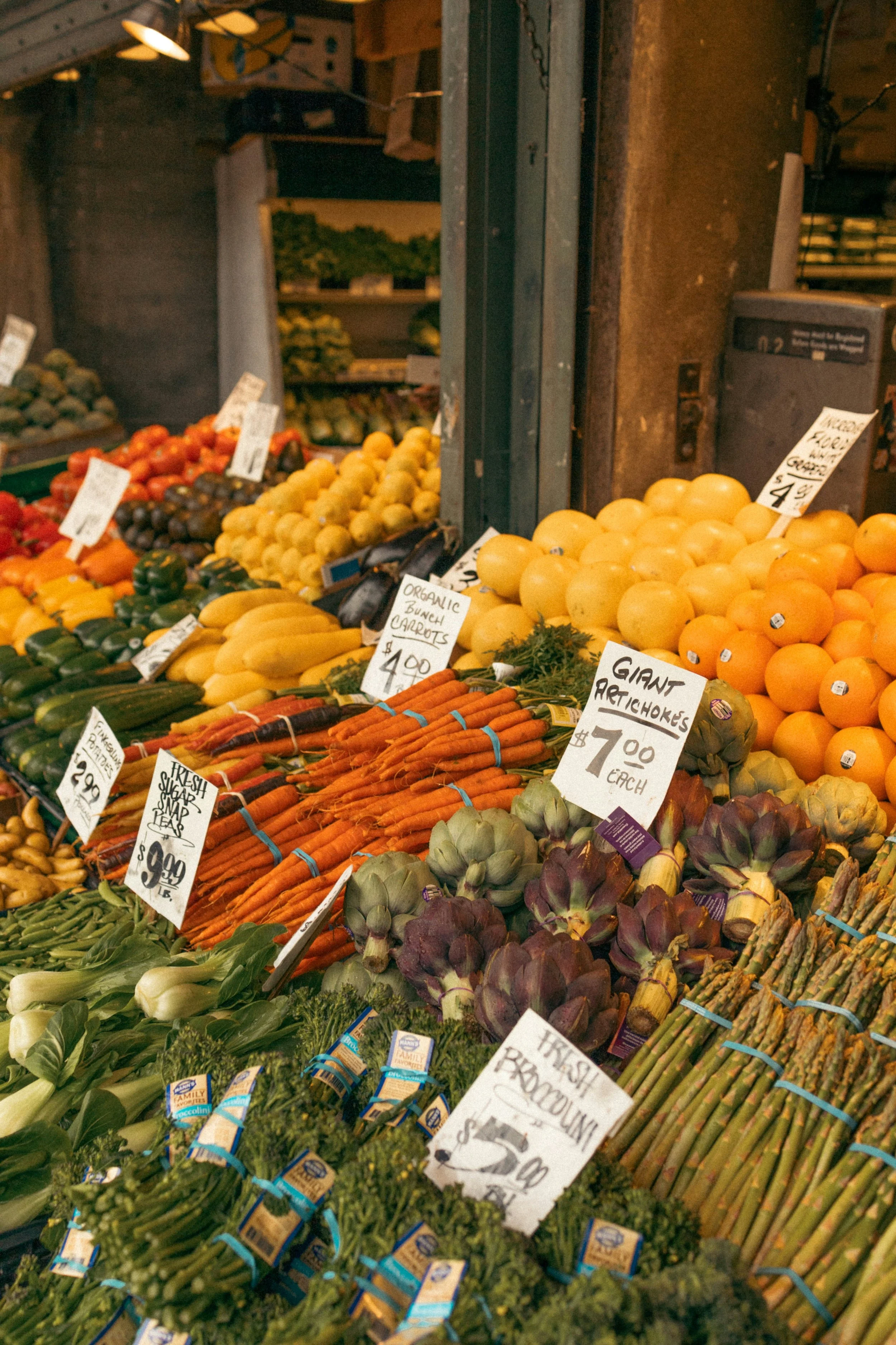 Display of fresh vegetables at a farmers market, including carrots, artichokes, asparagus, and leafy greens, with handwritten price tags.