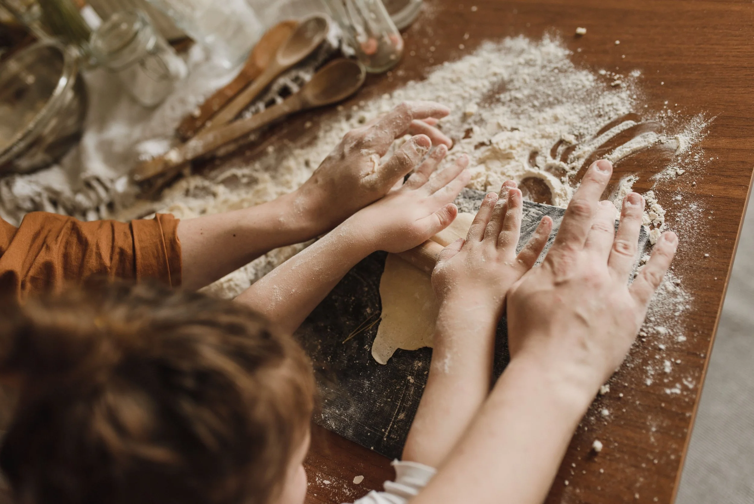 Multiple hands covered in flour working together to shape dough on a wooden surface, with baking utensils and ingredients visible in the background.